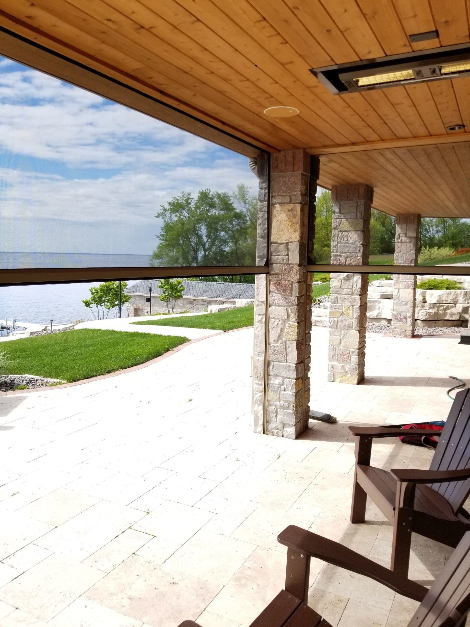 Outdoor patio with a partially lowered screen overlooking a water view.  A wooden ceiling and stone columns frame the space.