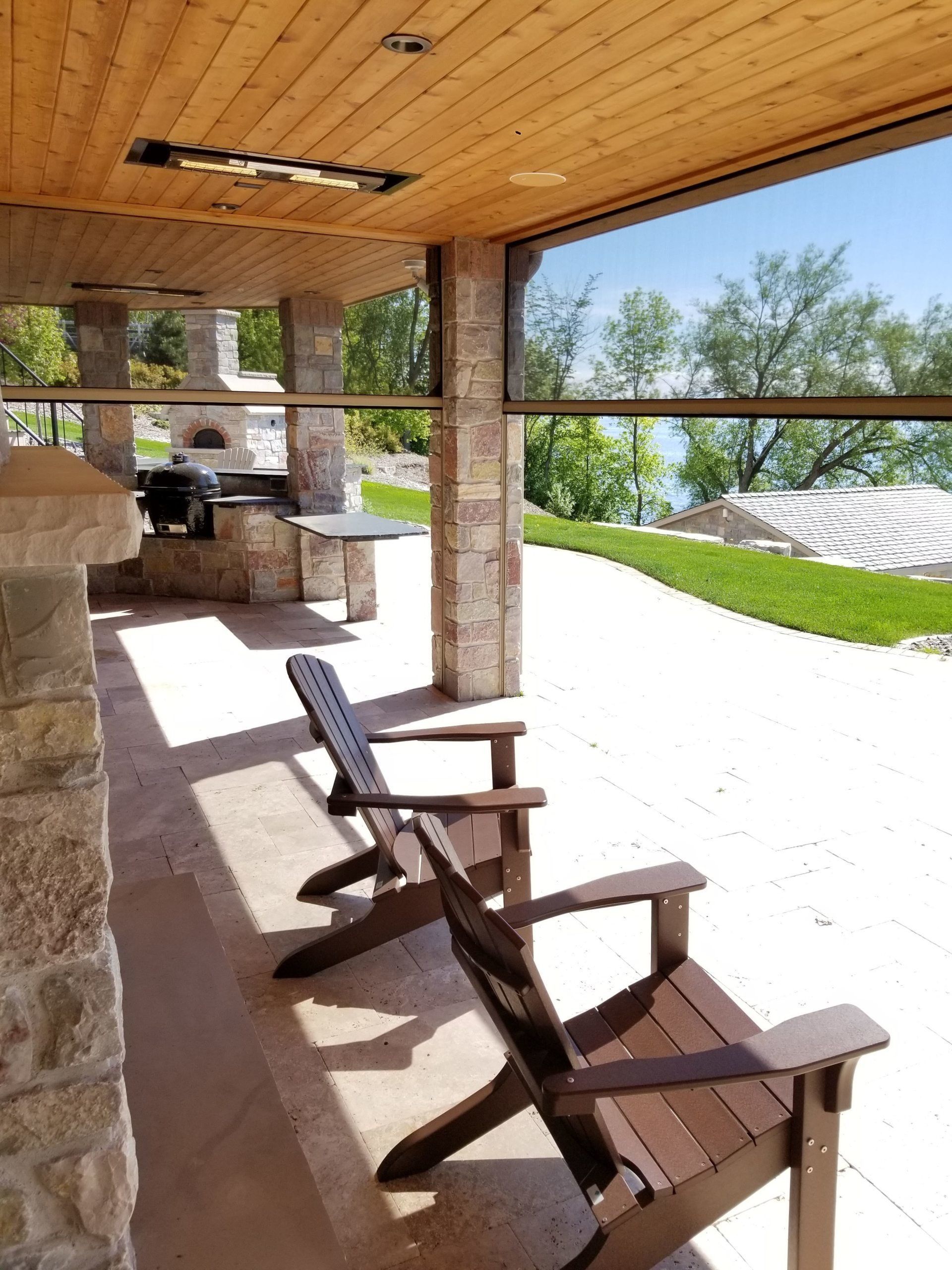 Two Adirondack chairs on a patio overlooking a waterfront view. The patio has stone columns, a wooden ceiling, and an outdoor kitchen.