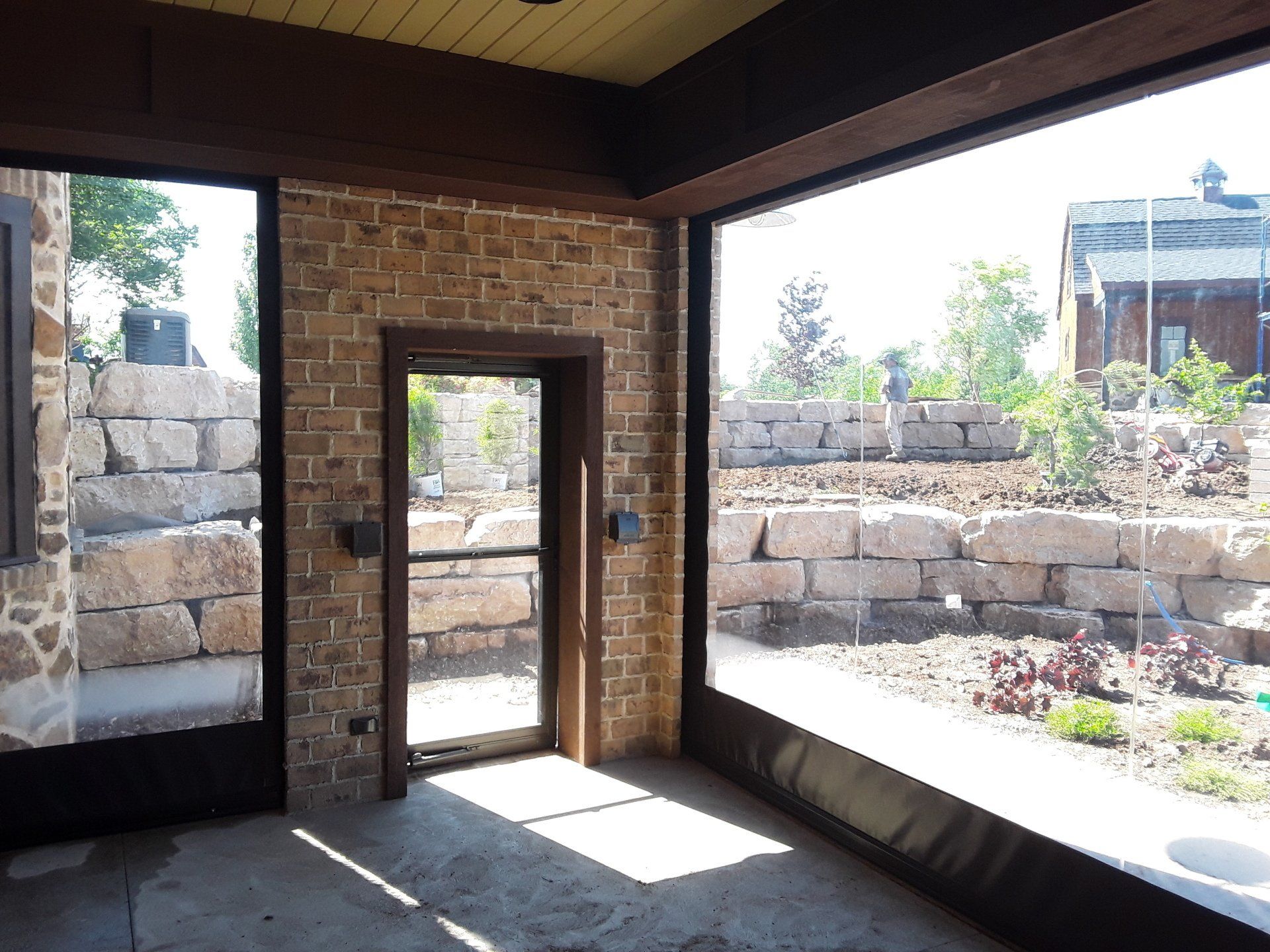 Interior view of a sunroom with stone walls and large windows. A screen door leads to an outdoor area with a stone retaining wall and landscaping.