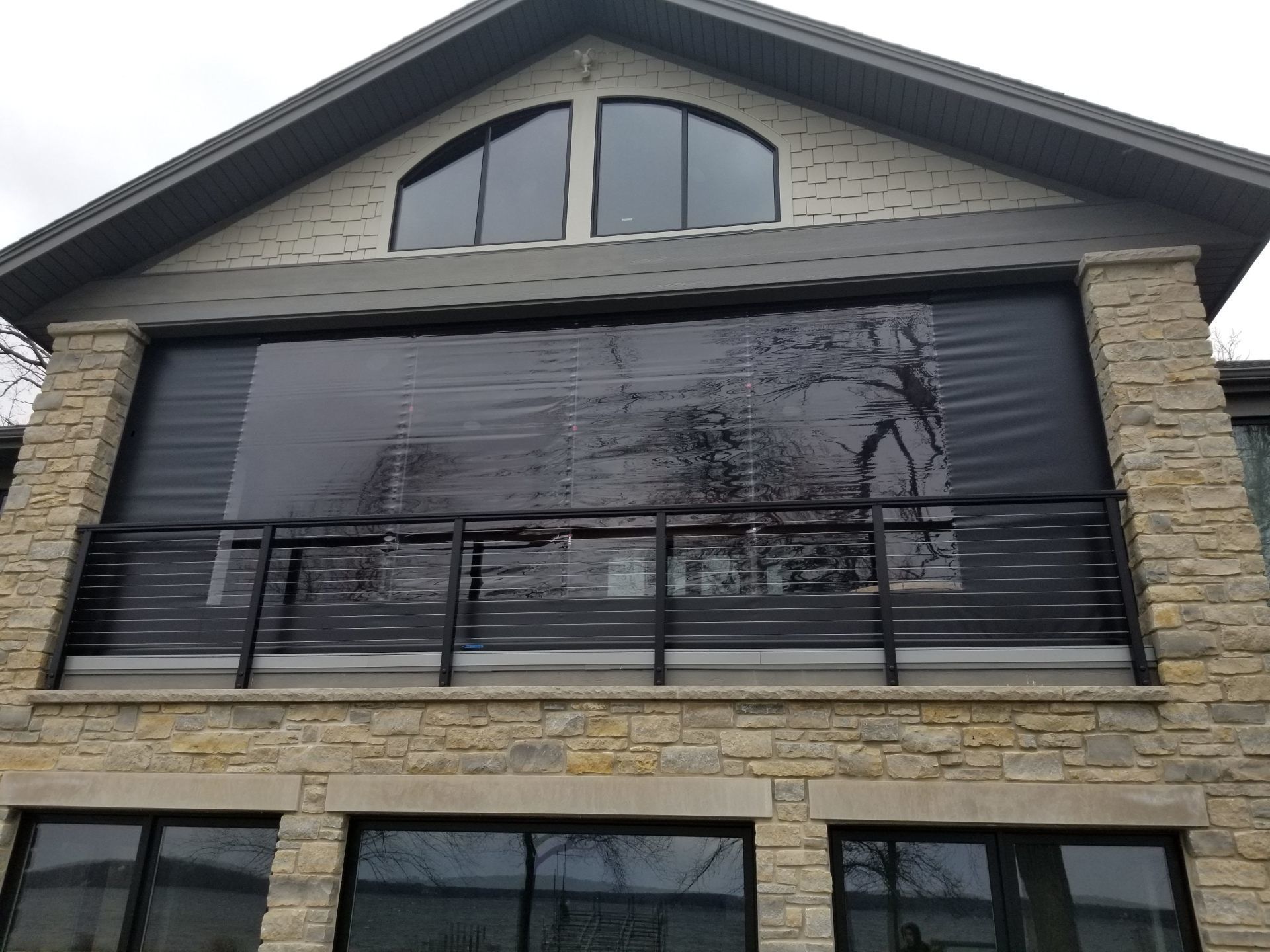 A two-story house with a stone exterior, featuring a balcony enclosed by black mesh screens and a black metal railing.
