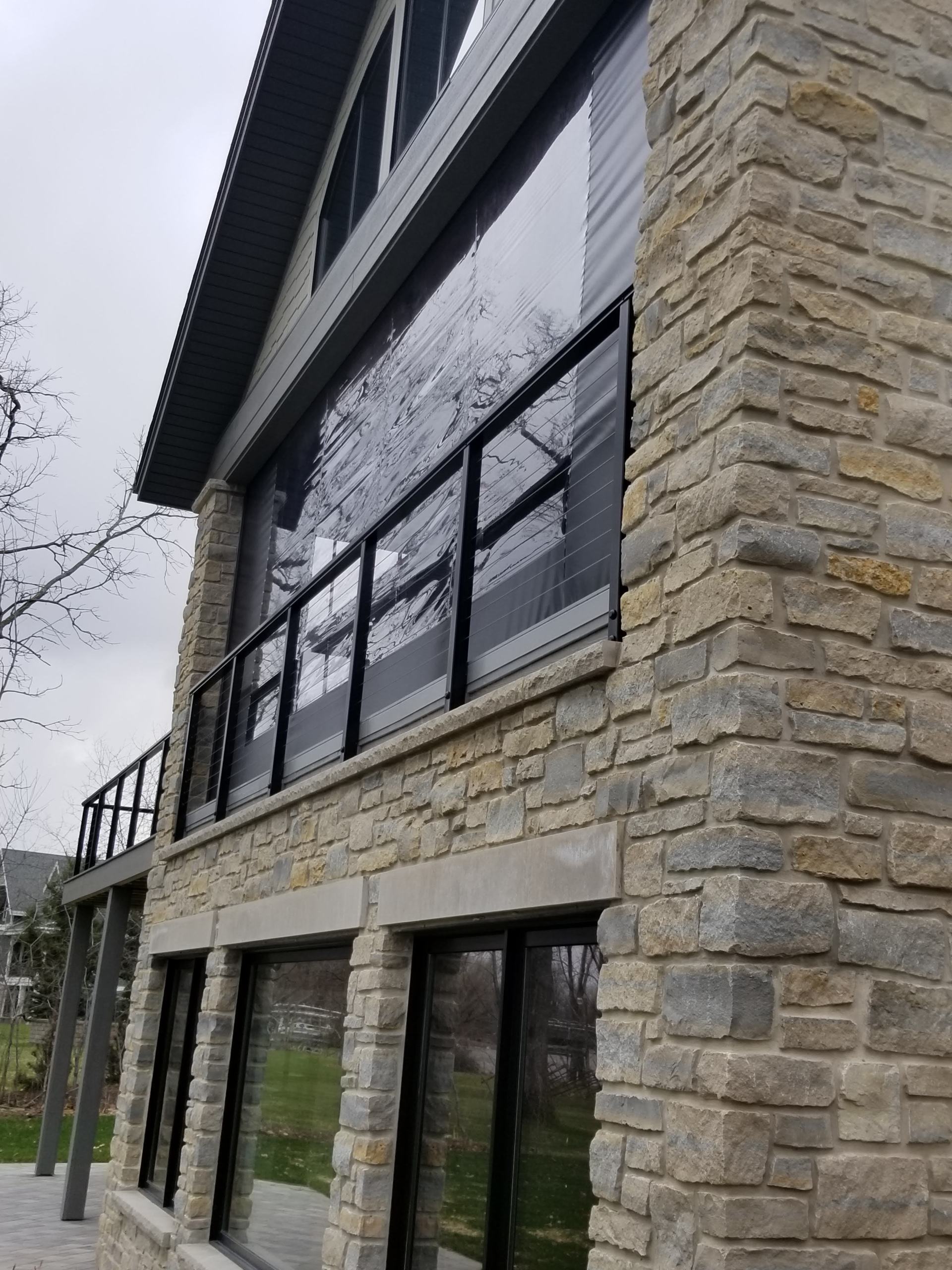 Stone house exterior with large windows, a black railing, and a dark roof. The sky is overcast.
