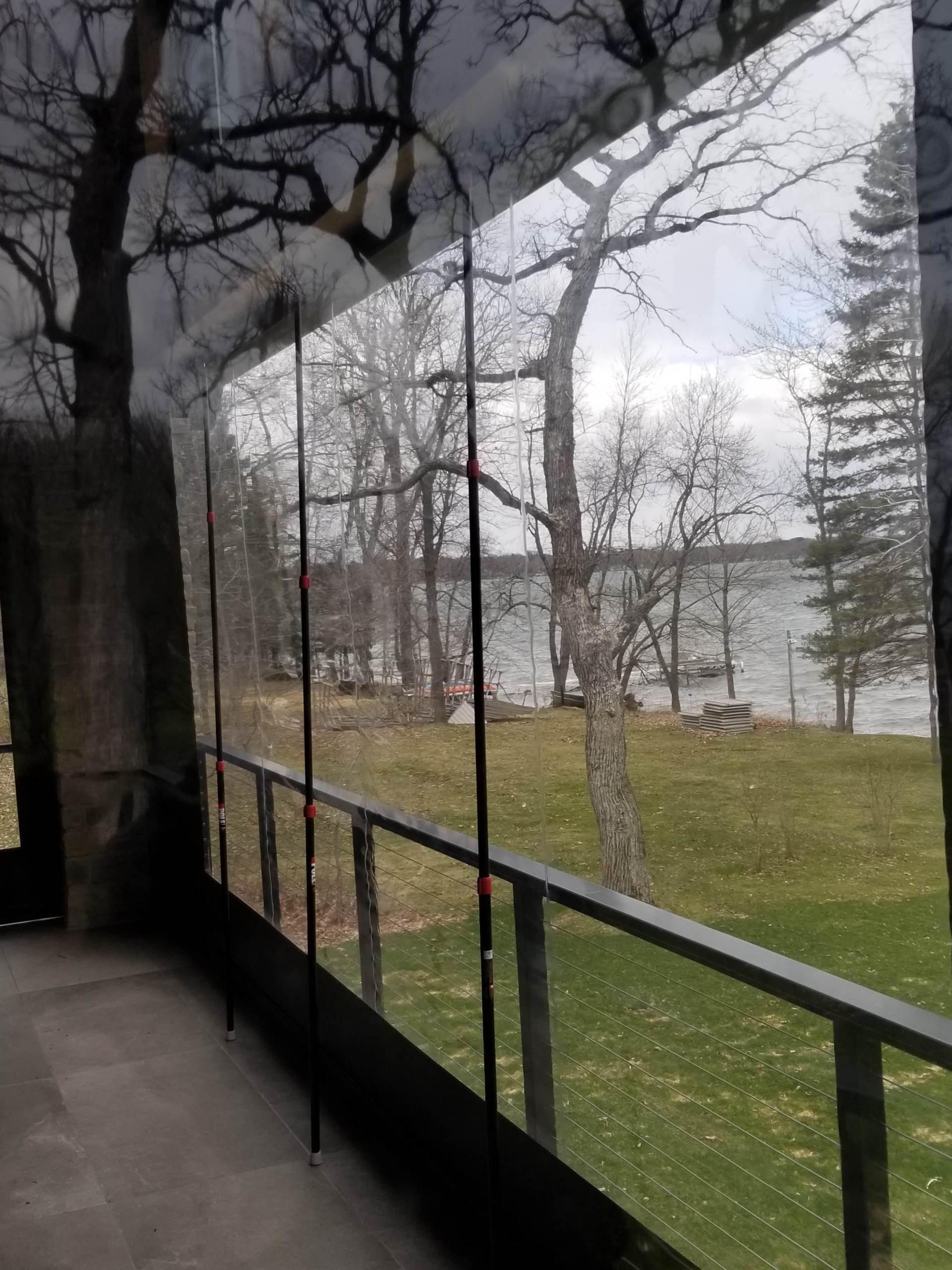 A clear, flexible vinyl enclosure on a porch with a view of a lake and trees. The enclosure is dark-framed and partially rolled up.