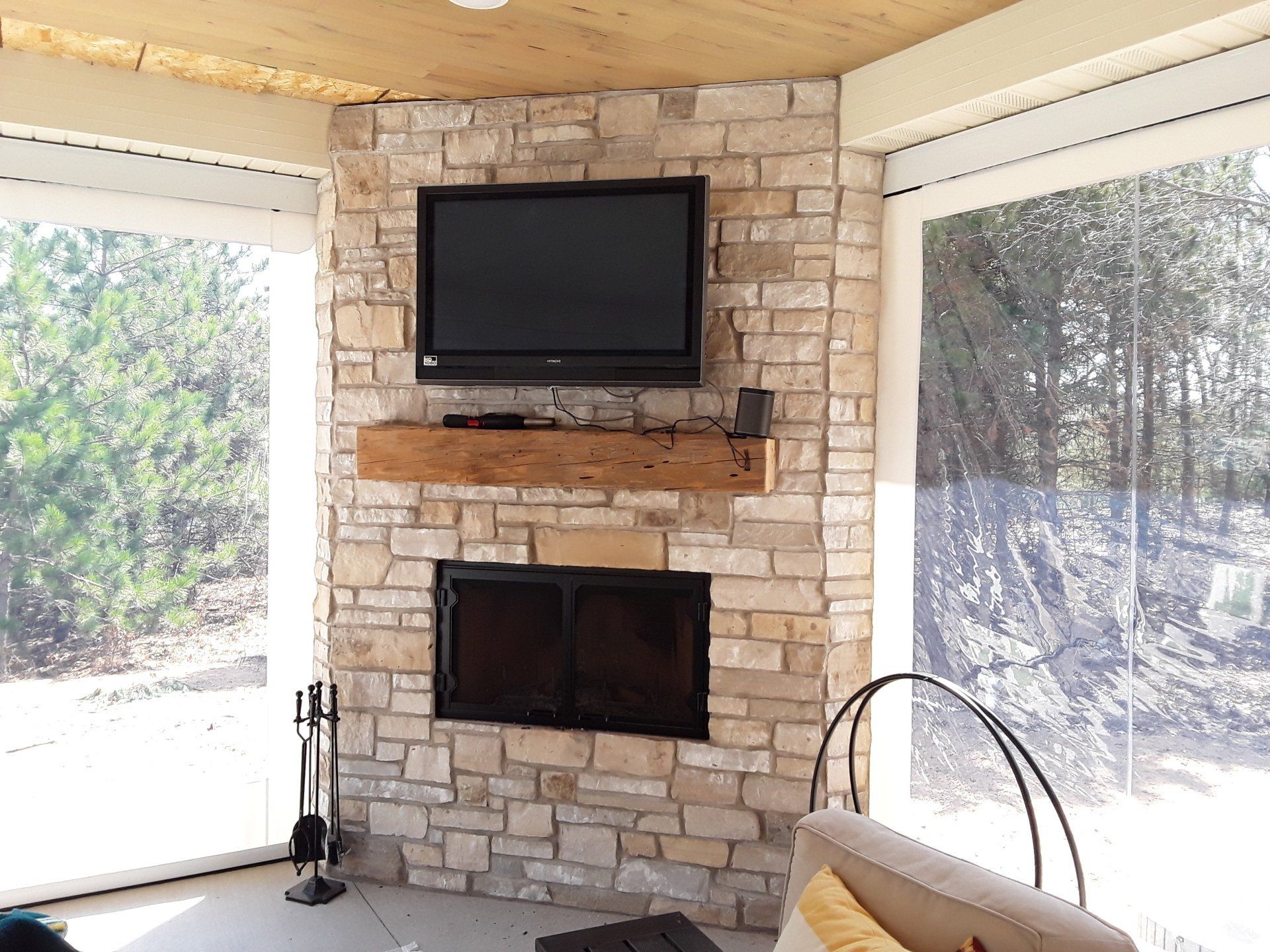 A stone fireplace with a TV and wooden mantle in an enclosed porch. Large windows provide a view of trees.