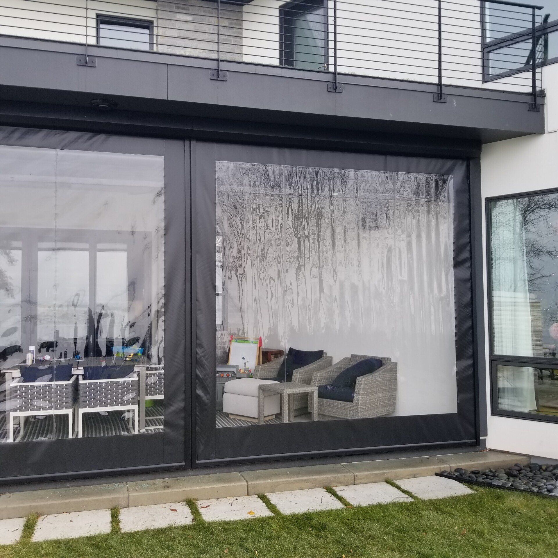 Black-framed clear vinyl patio enclosures installed on a modern home with outdoor furniture visible inside.