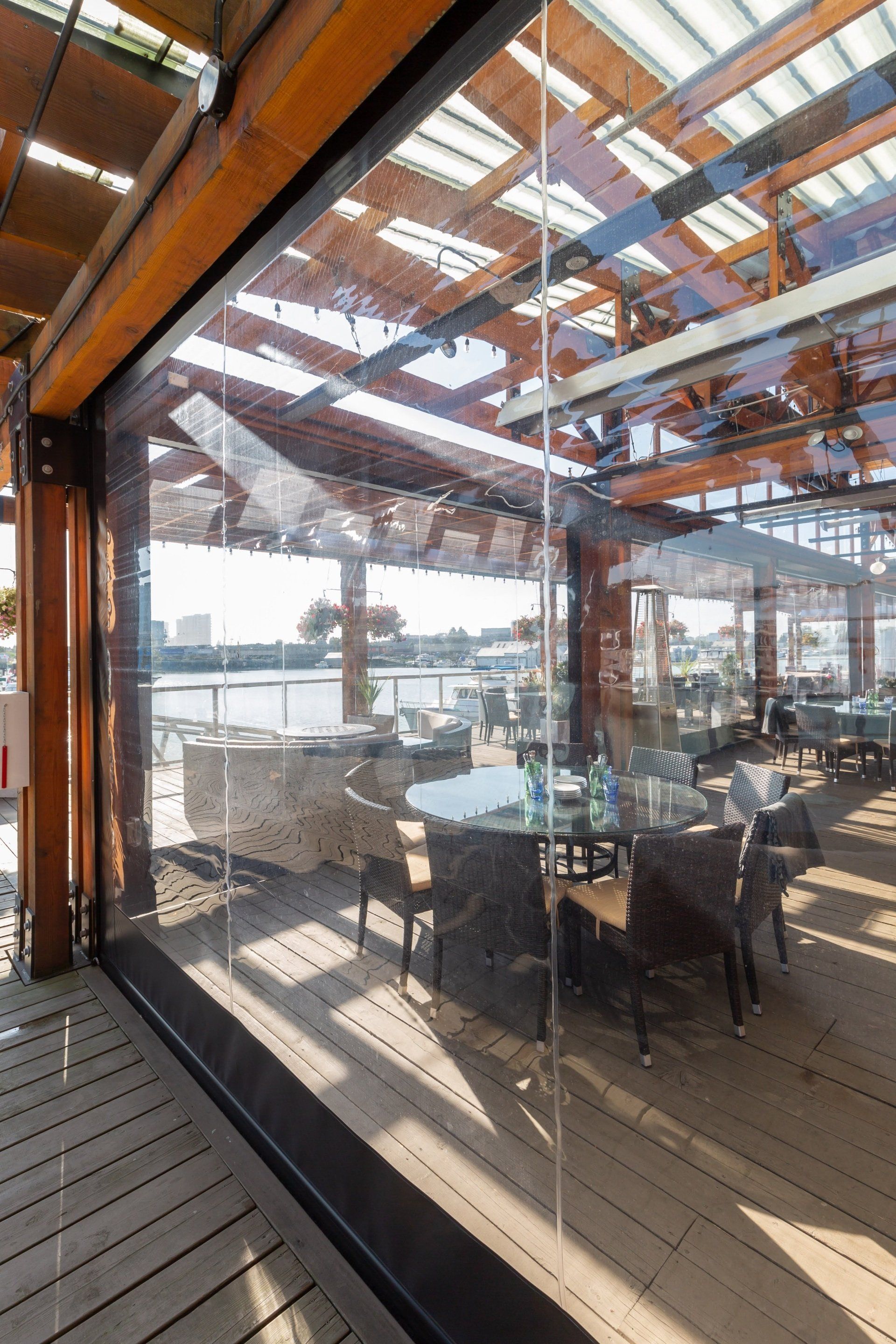 A clear, partially enclosed outdoor dining area with tables and chairs. Natural wood and glass structure.