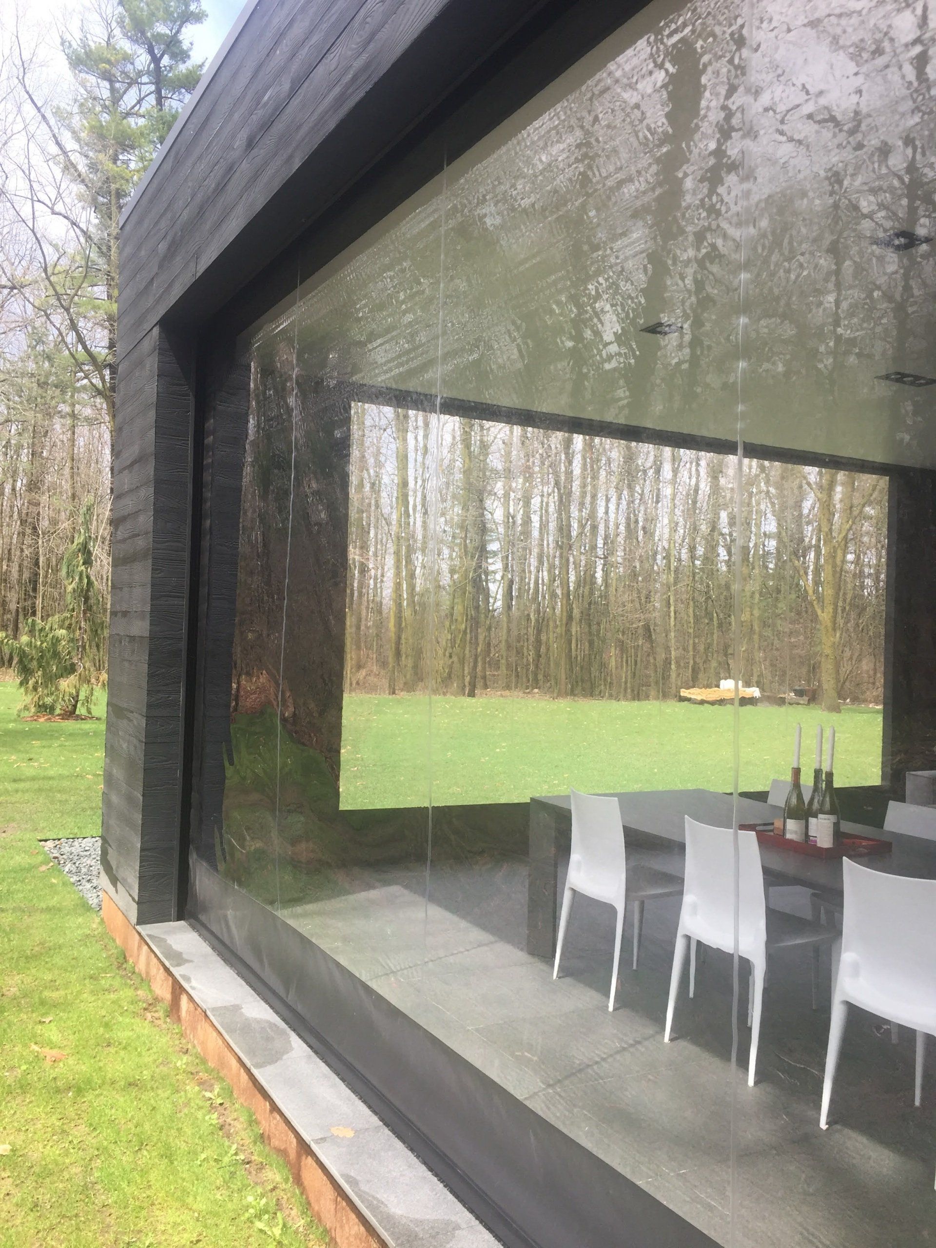 Modern dining space with large glass windows reflecting a wooded landscape and a table with white chairs.