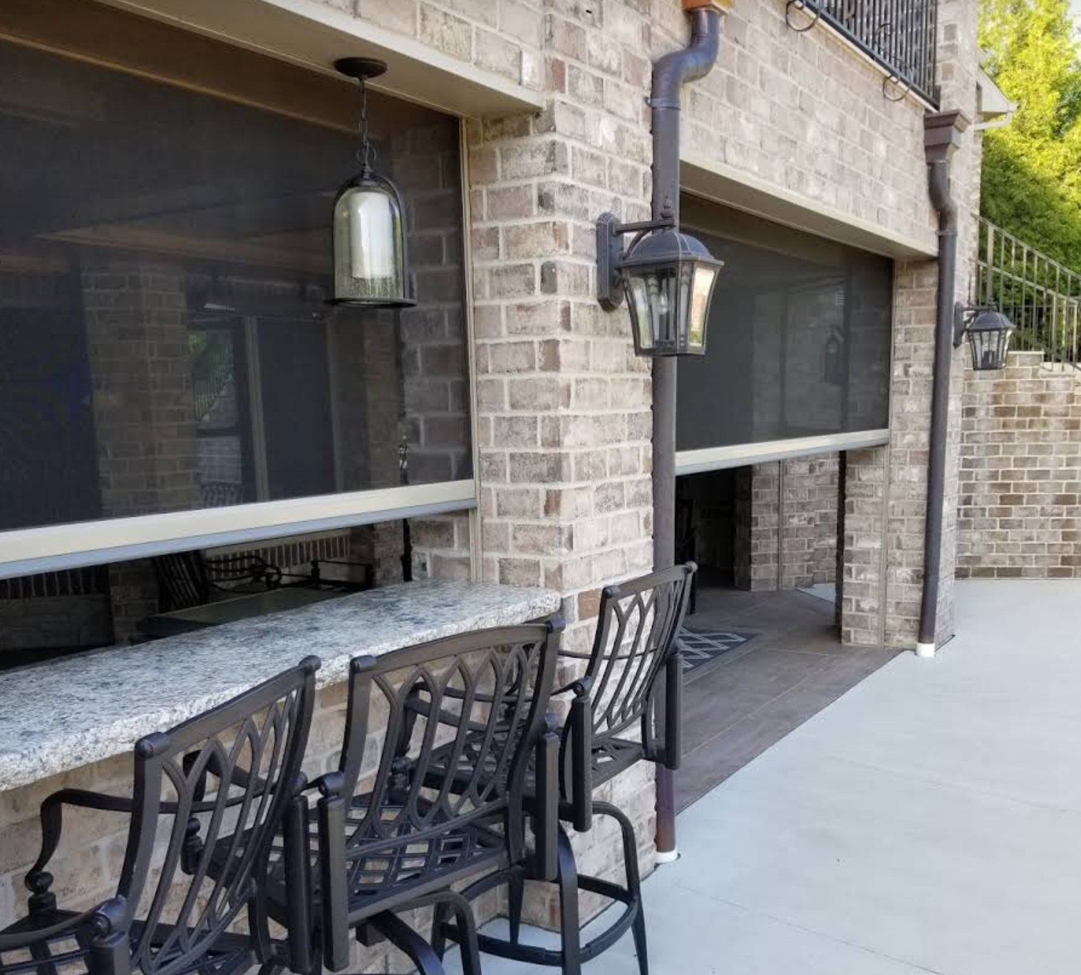 Outdoor bar area with retractable screens, brick columns, and metal bar stools. Two pendant lights and two wall-mounted lanterns are visible.