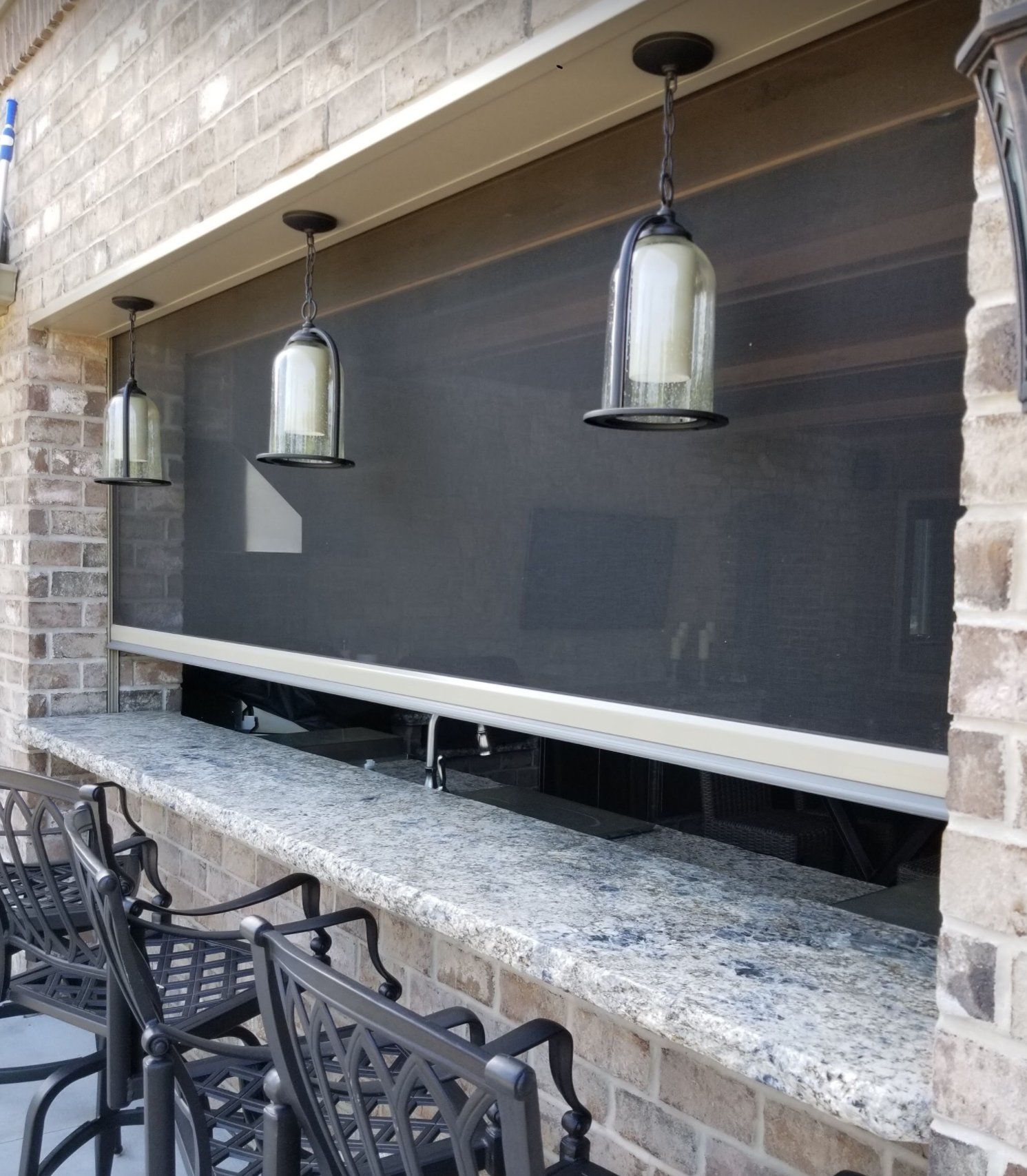 Outdoor bar with granite countertop and black metal chairs. A roll-down screen and hanging lights are above.