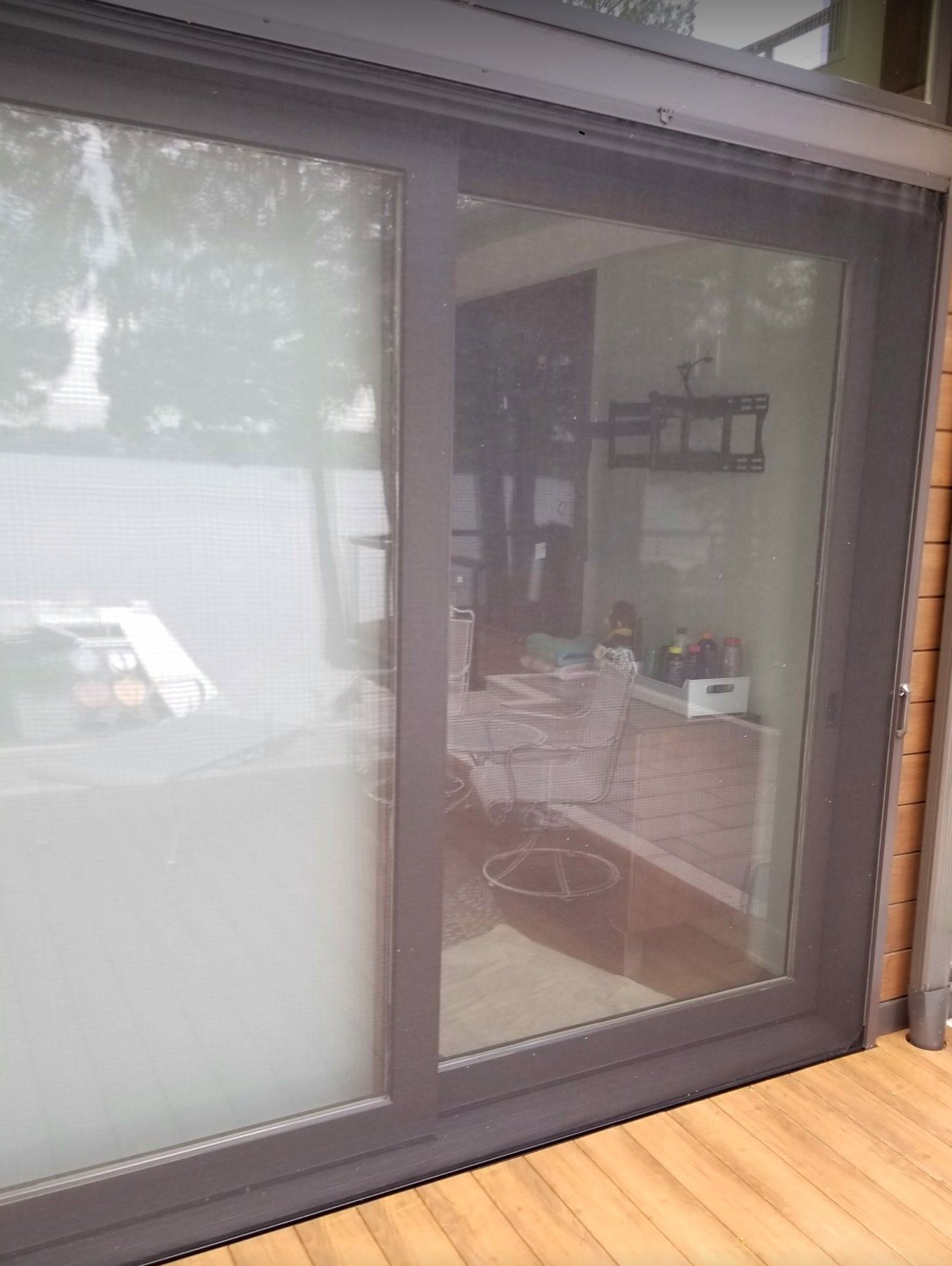 Sliding glass door with a dark gray frame, reflecting an interior room with furniture and an exterior deck.
