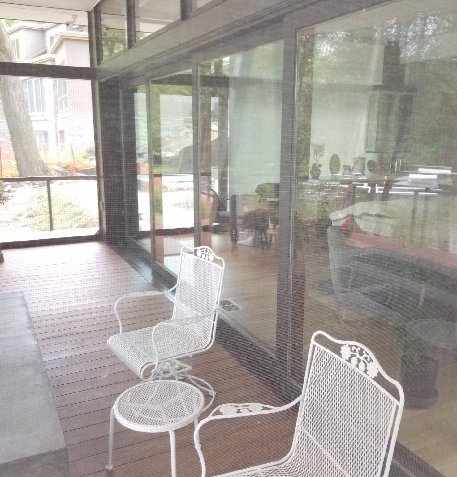 Two white wrought-iron chairs and a small table on a wooden porch with floor-to-ceiling glass windows.