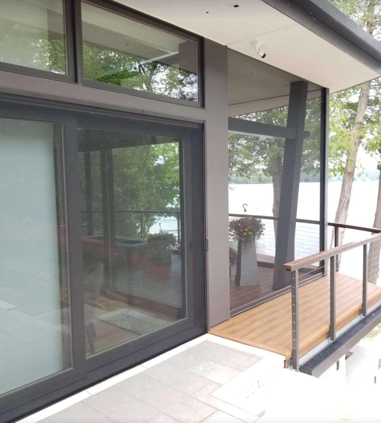 Modern lakeside house with large sliding glass doors, a deck, and a view of trees and water. The house has gray and dark-colored elements.
