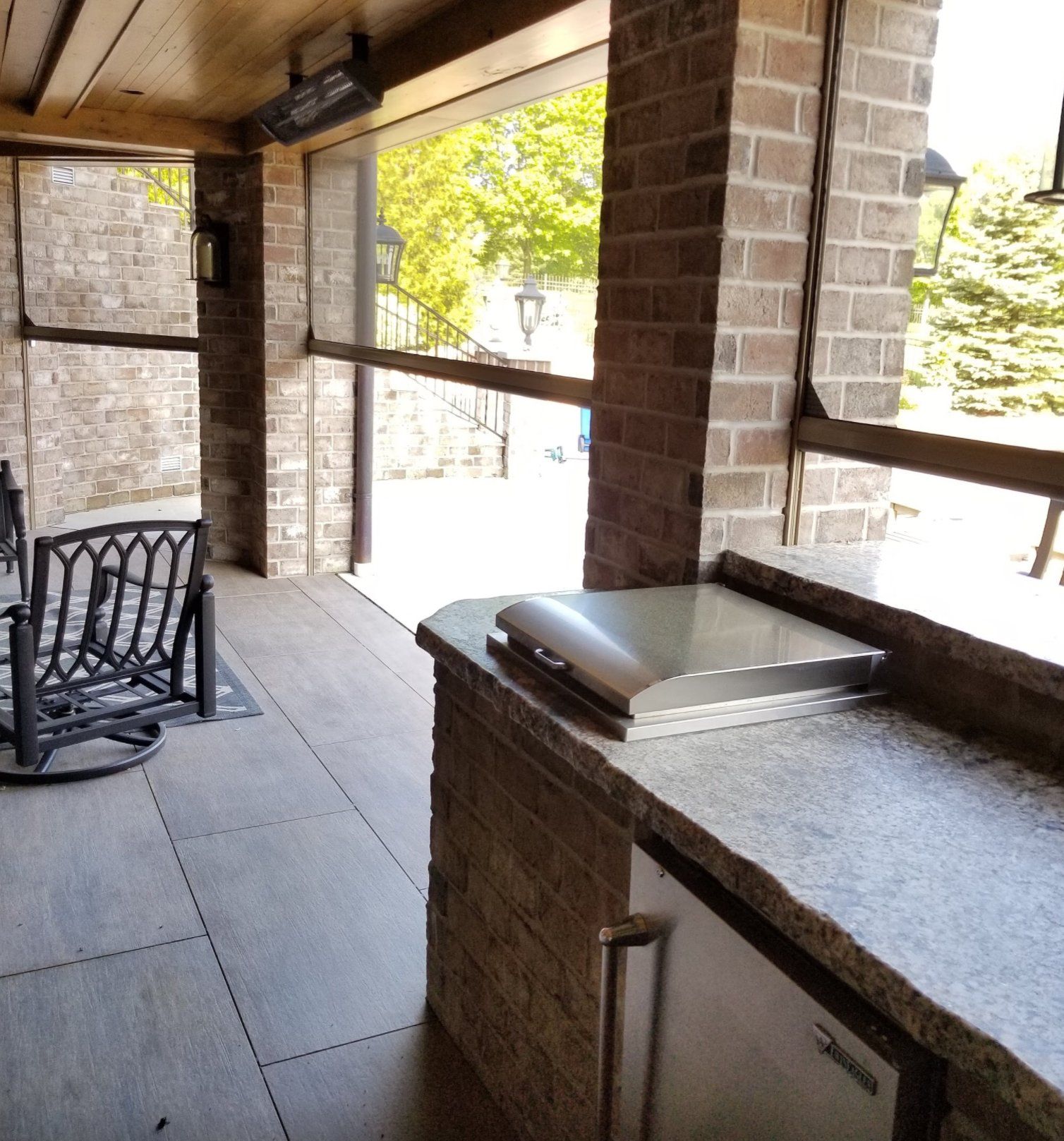 Outdoor kitchen with brick columns, a grill, and a counter. Light-colored floor and screens visible, with a view of greenery outside.