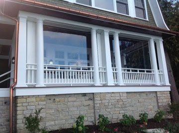 White porch with large windows, supported by columns, over a stone foundation. Small bushes are in the foreground.