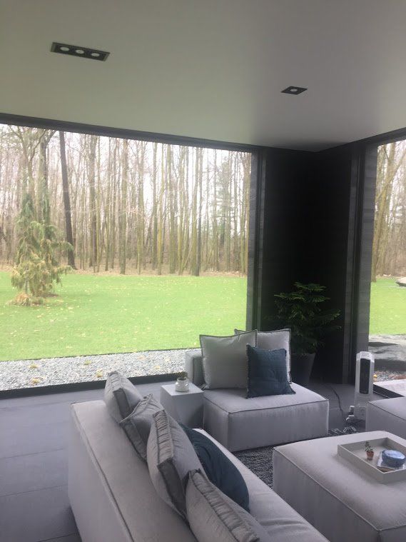 Modern living room with large windows overlooking a green lawn and trees. White couches with grey and blue pillows.