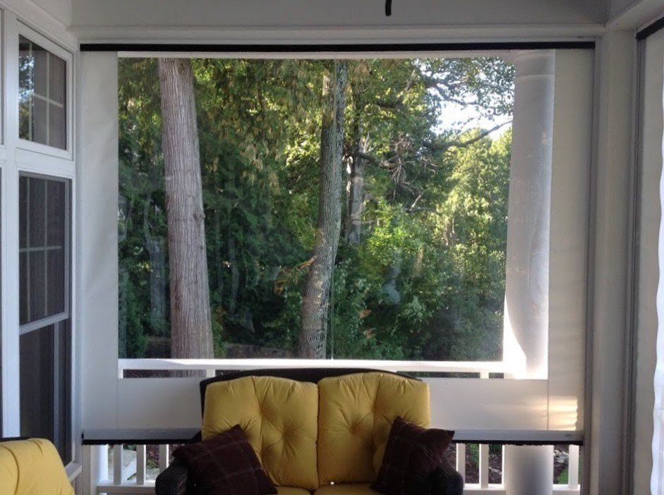 Sunroom with a view of a lush forest, featuring beige roller shades over a two-person couch with yellow cushions.