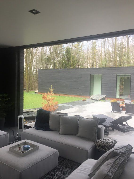 Interior view looking out onto a modern patio with a grey sofa, overlooking a grassy area and a contemporary building nestled in a wooded area.