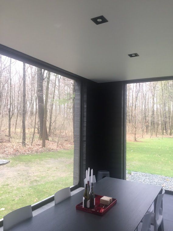 Modern dining area with large windows overlooking a wooded area. Dark gray table and chairs. White ceiling with recessed lighting.