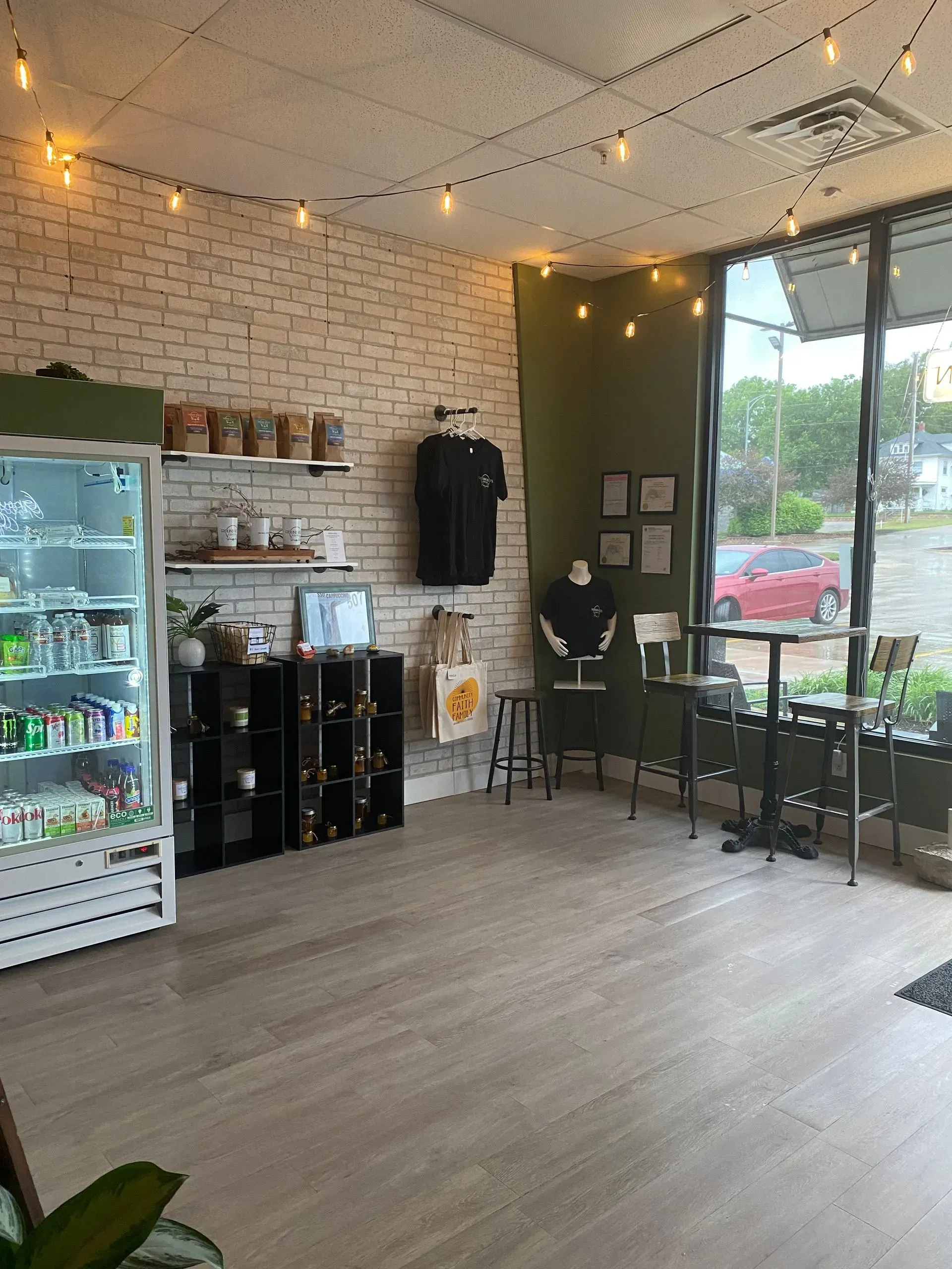 Interior of a shop with a fridge, display shelves, brick wall, string lights, and tables with seating by a window.