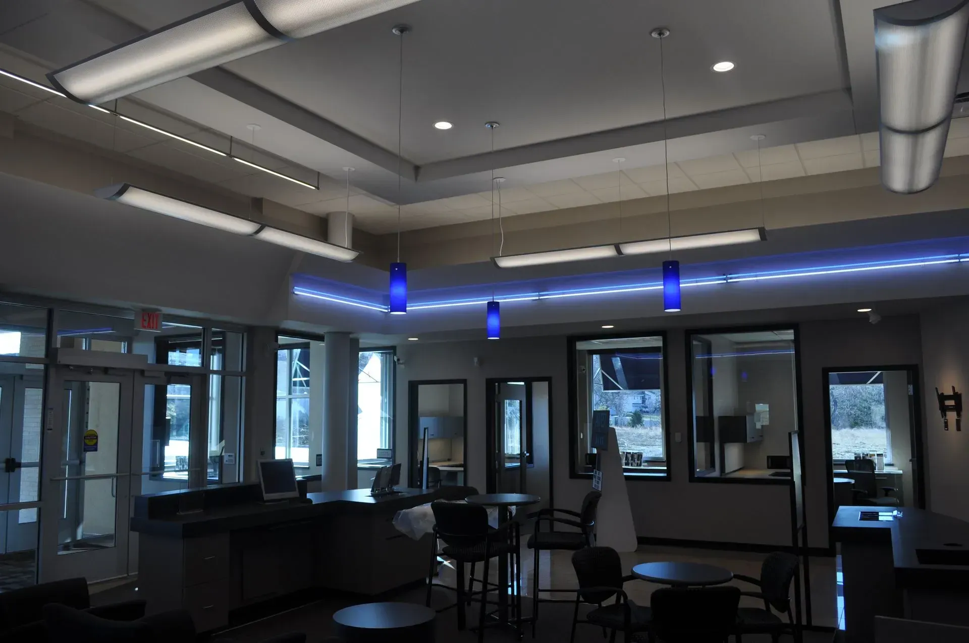 Modern, well-lit interior with glass doors and windows, tables, chairs, and hanging blue lights.