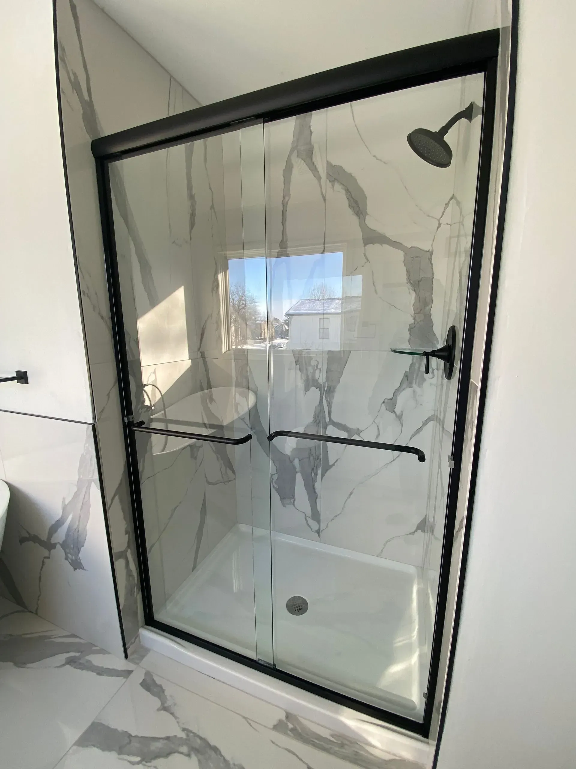 Black-framed sliding glass shower door in a bathroom with marble-look tiled walls and white shower base.