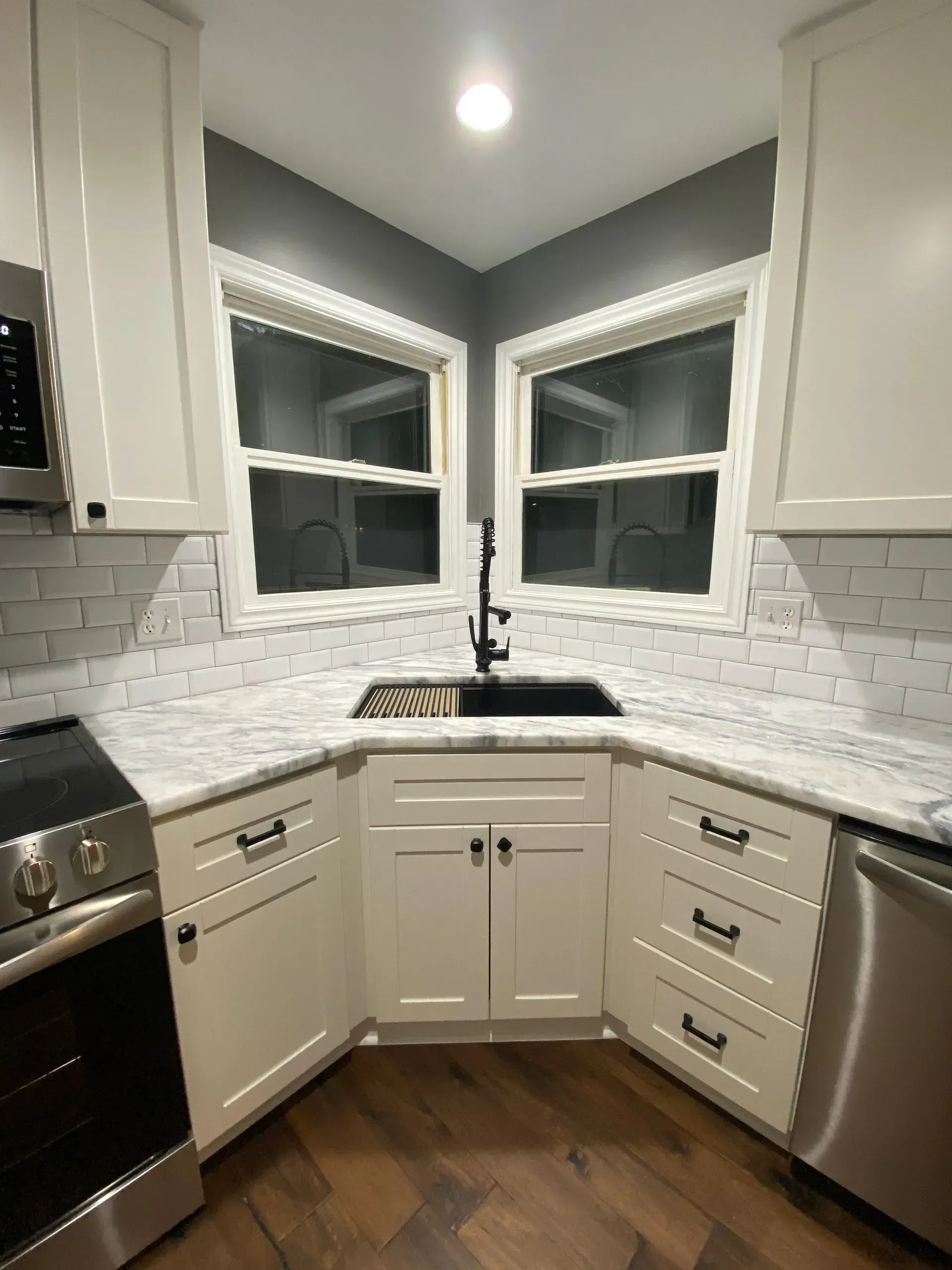 White kitchen cabinets surround a sink in a corner, with a black faucet. Windows and gray walls are above.