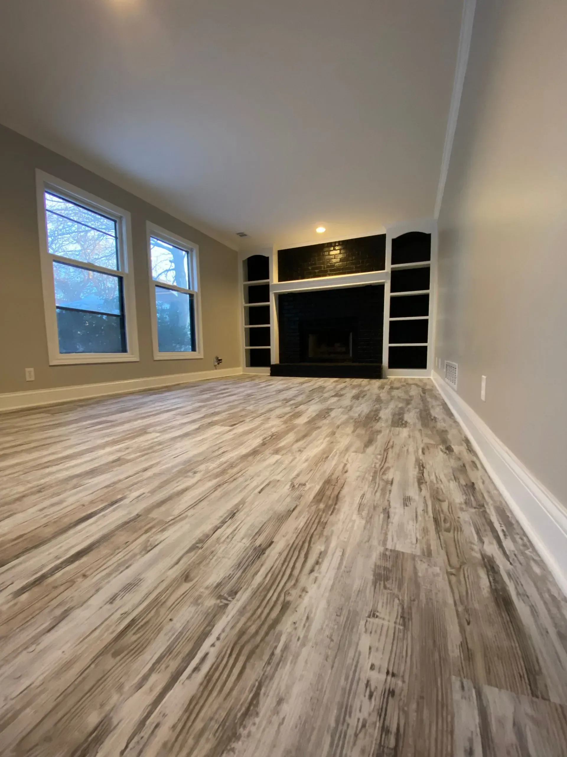 Empty living room with light wood-look flooring, fireplace, built-in shelves, and windows.