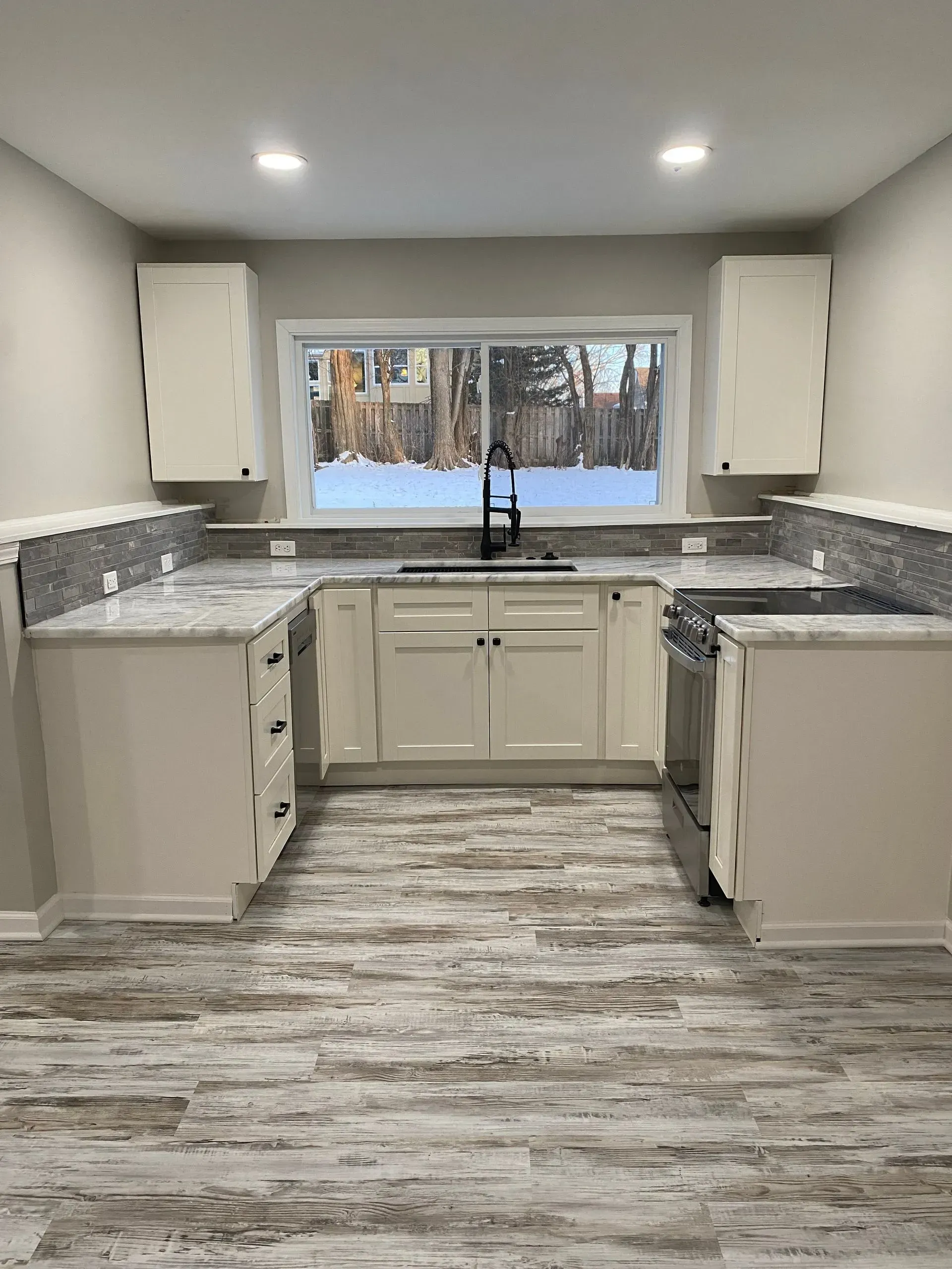 U-shaped kitchen with white cabinets, gray countertops, and wood-look flooring. A window overlooks a snowy outdoor scene.