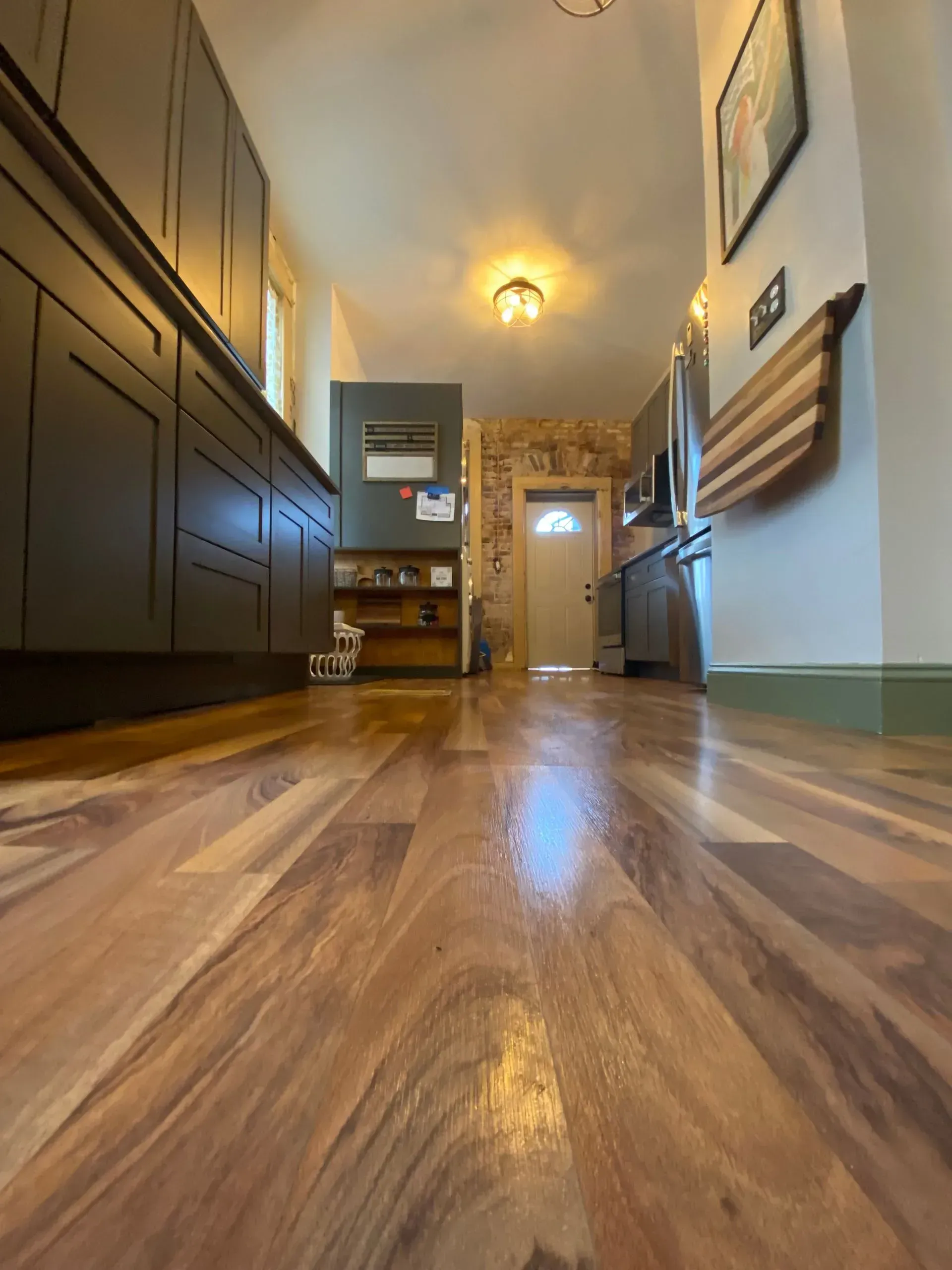 Low-angle view of a narrow kitchen with wooden floors, dark cabinets, and a doorway at the end.