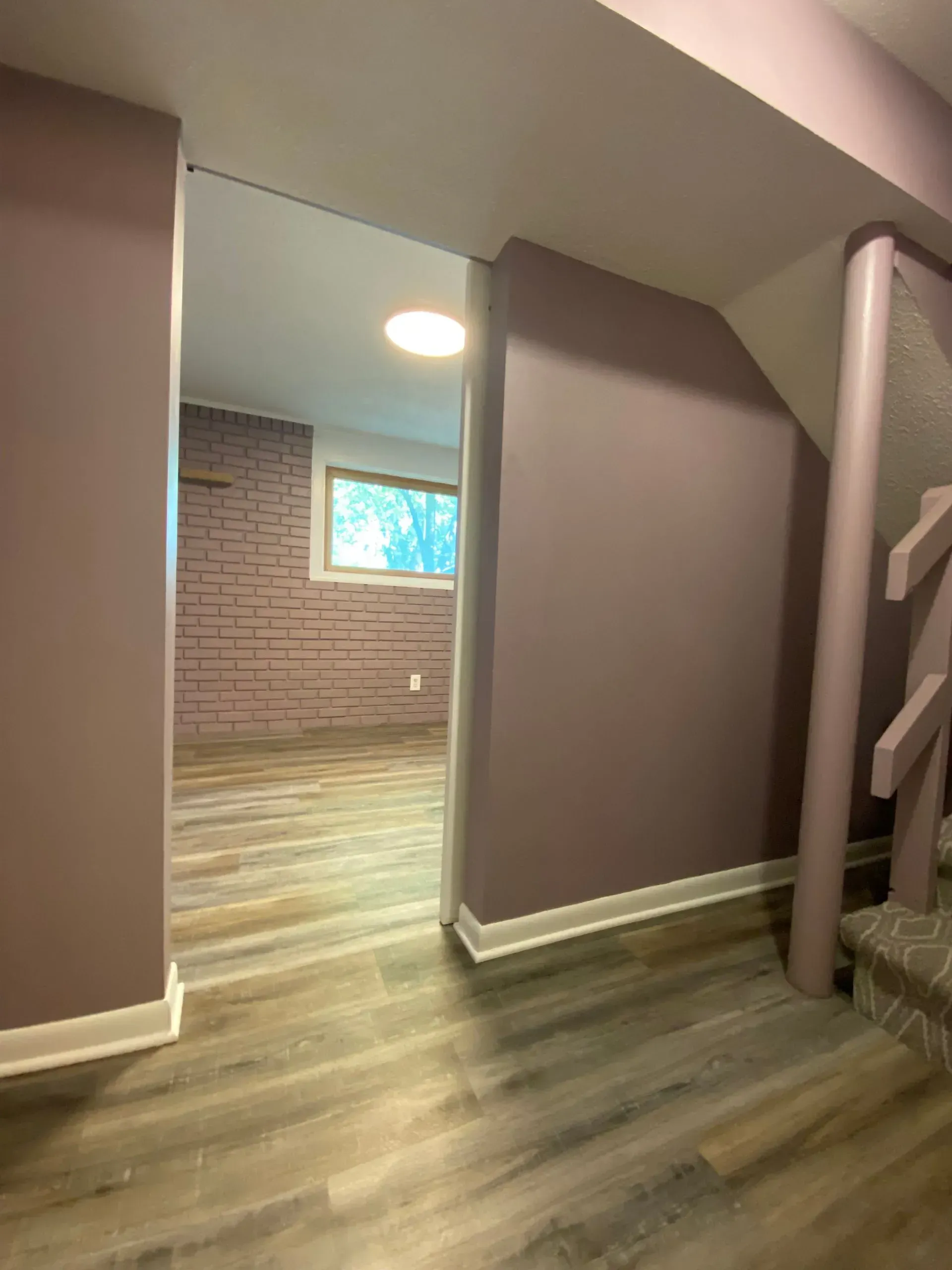 Interior view of a purple-painted basement hallway, leading to a room with a window and brick wall.