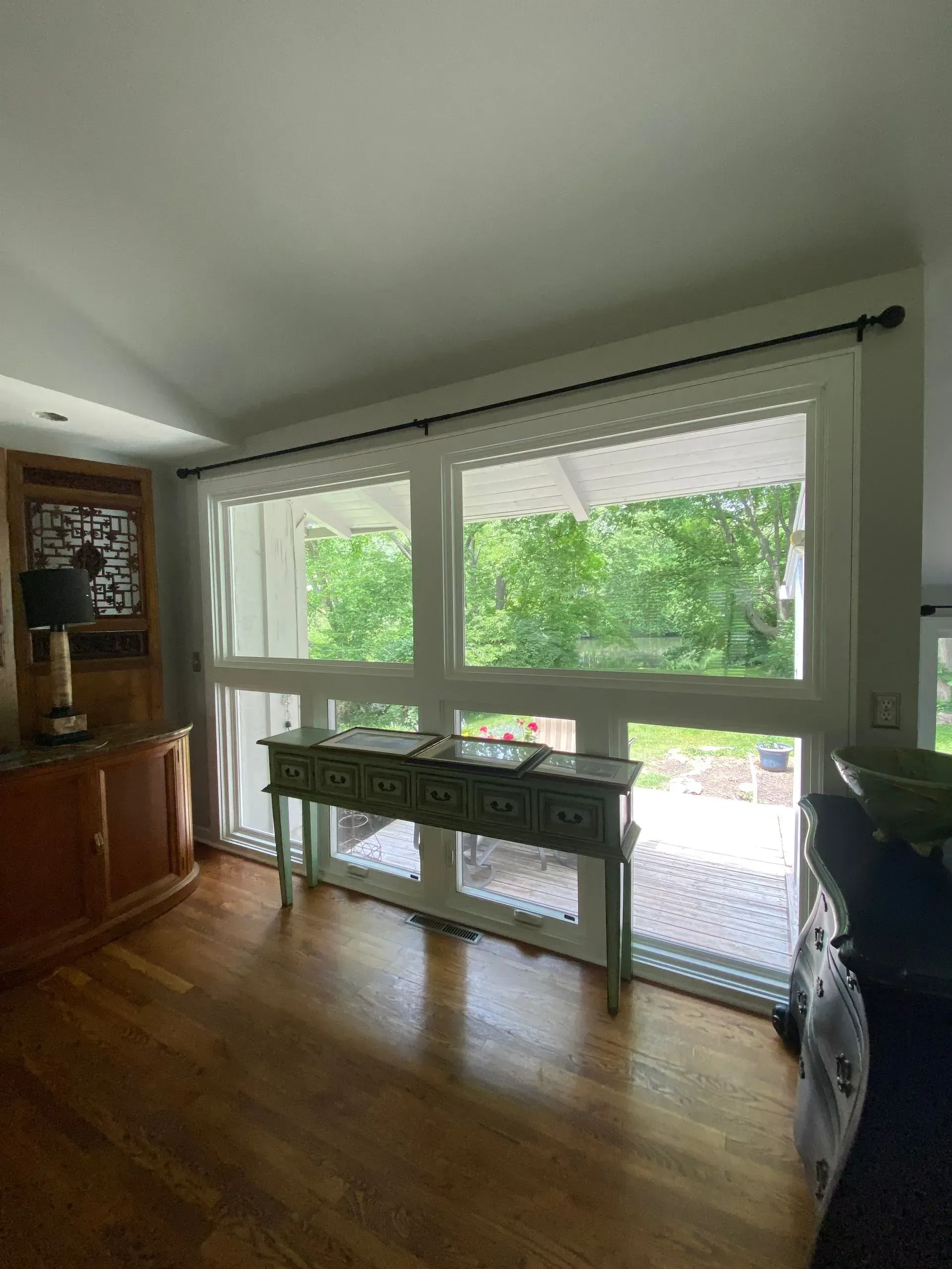 Room with wooden floor, large windows overlooking trees, and a decorative console table.