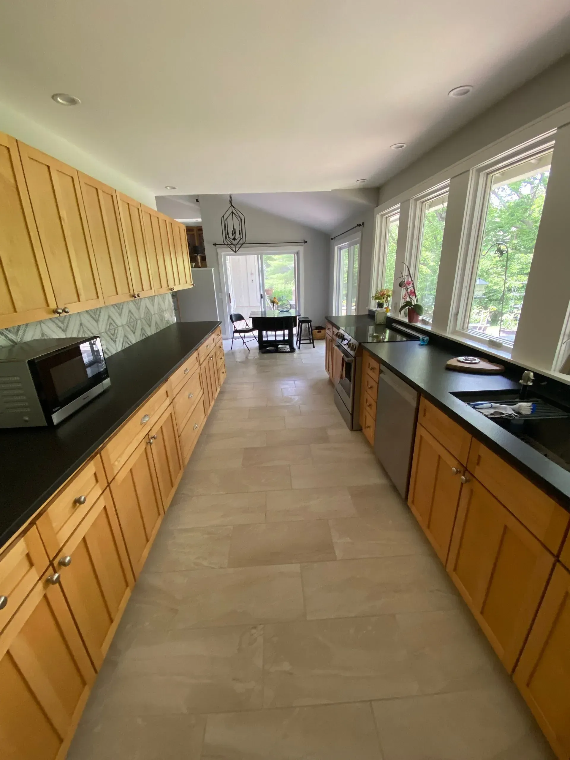 Long, narrow kitchen with light wood cabinets, black countertops, and tile floor.
