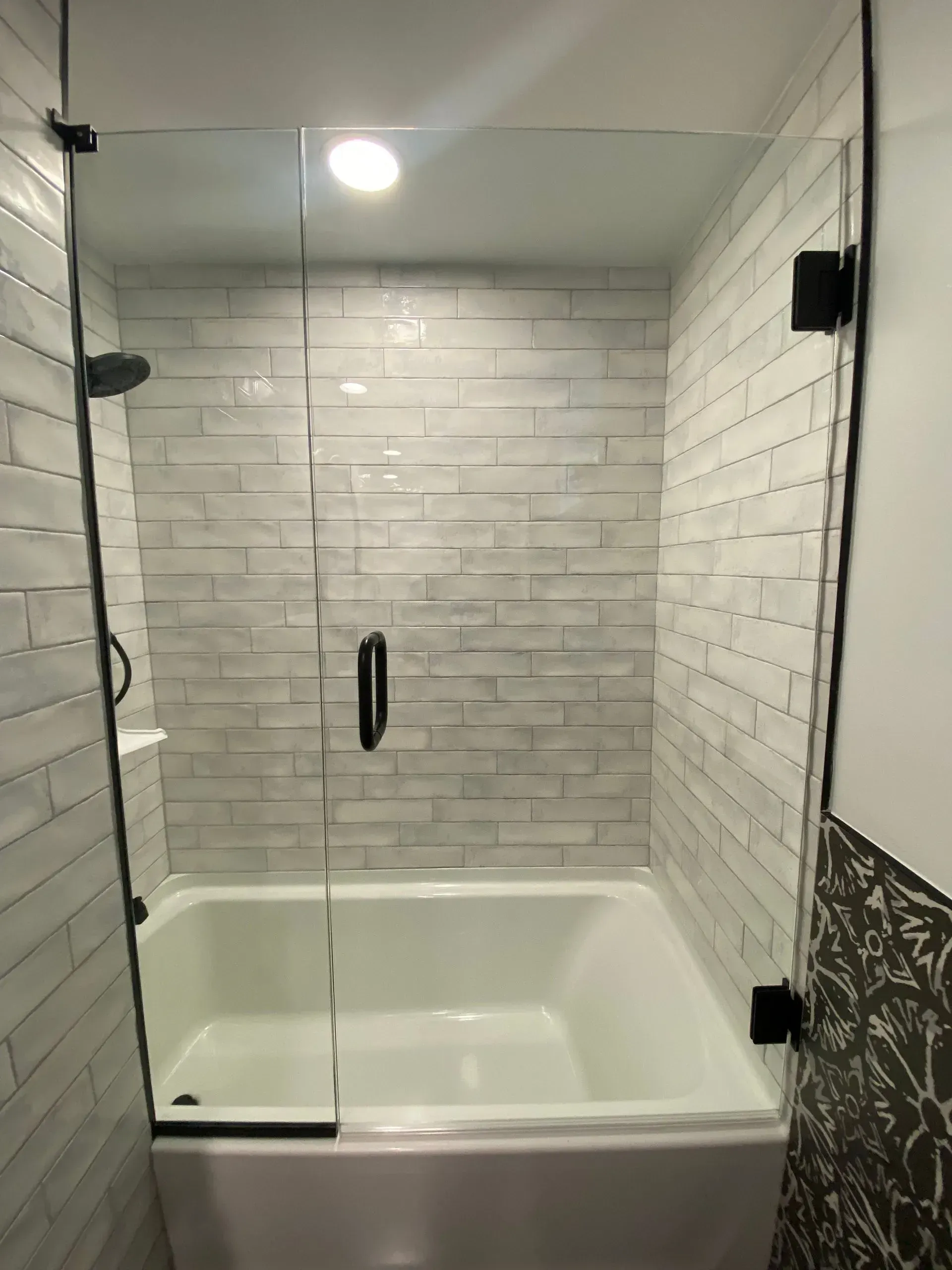 Bathtub with clear glass shower door and black hardware. White rectangular tile lines the wall.