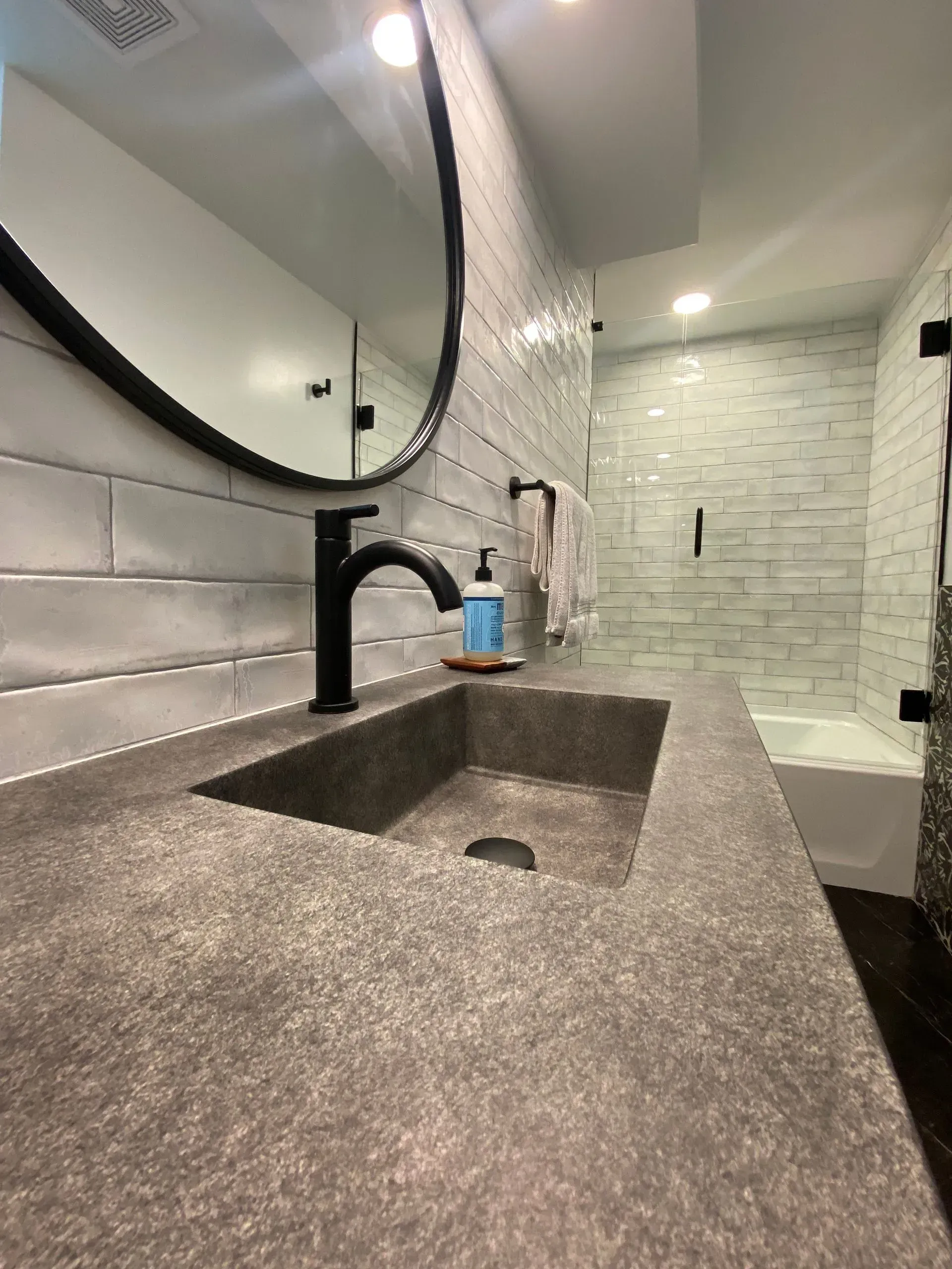 Bathroom with gray countertop, black faucet, round mirror, white tile, and glass shower.