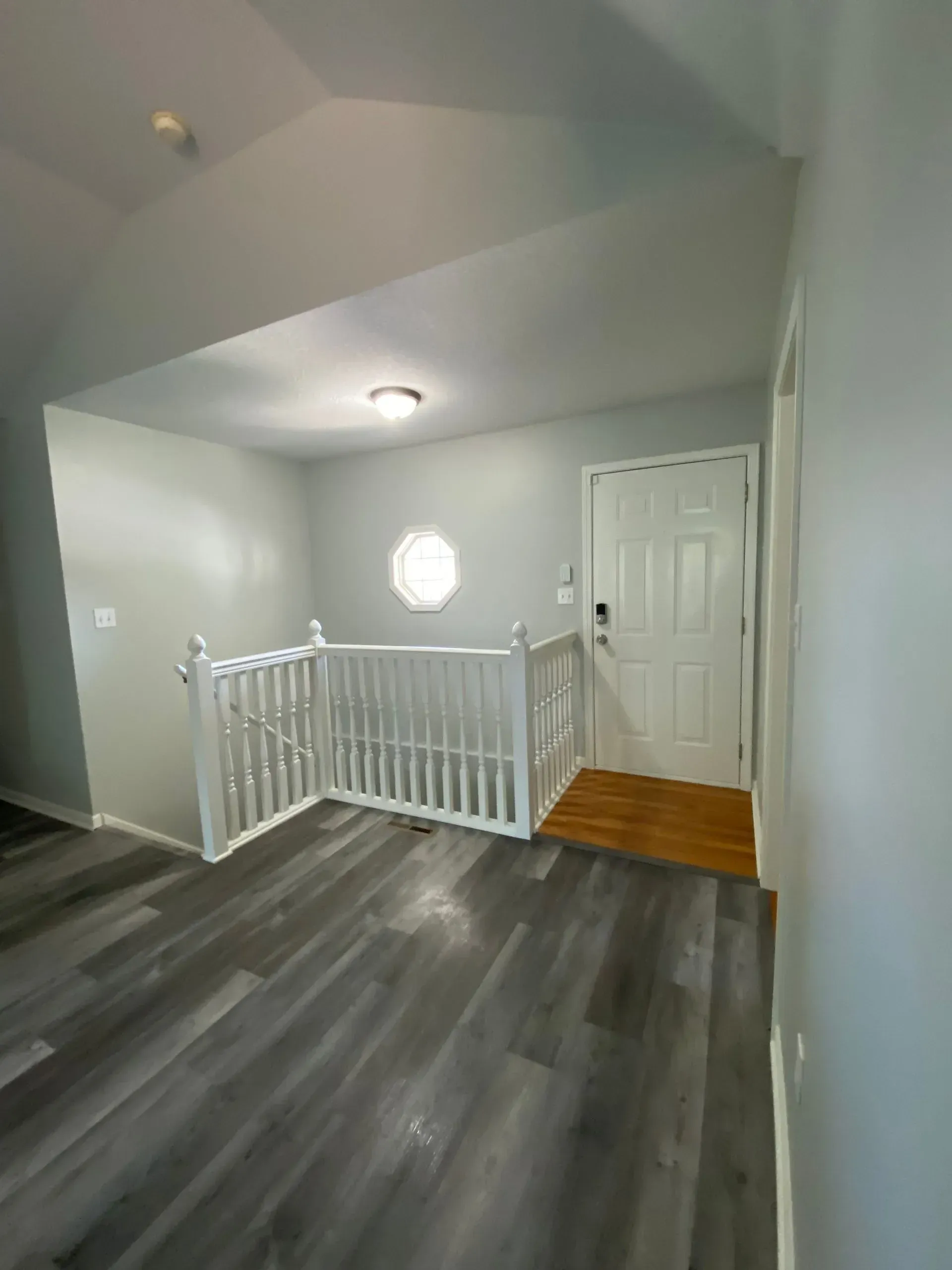 Interior of a home: gray flooring, light gray walls, white banister, door, circular window, and a light fixture.