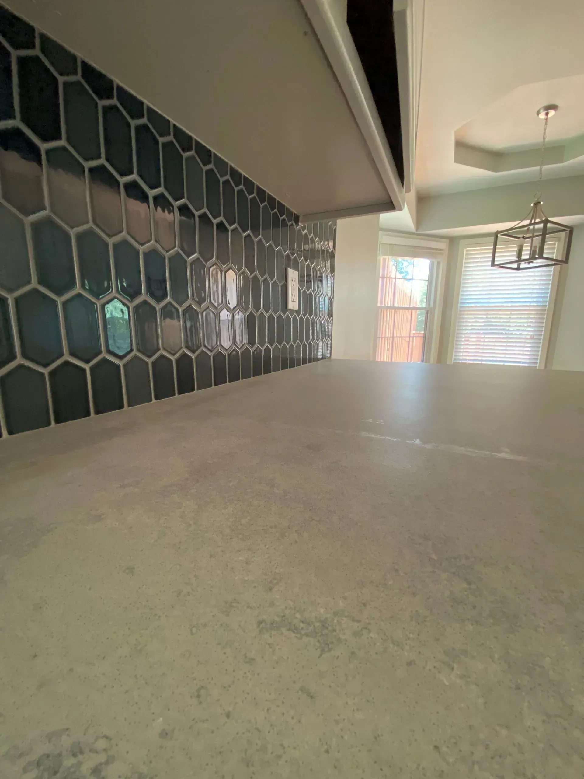 Gray countertop with dark hexagon-tiled backsplash, and a dining area with a chandelier.