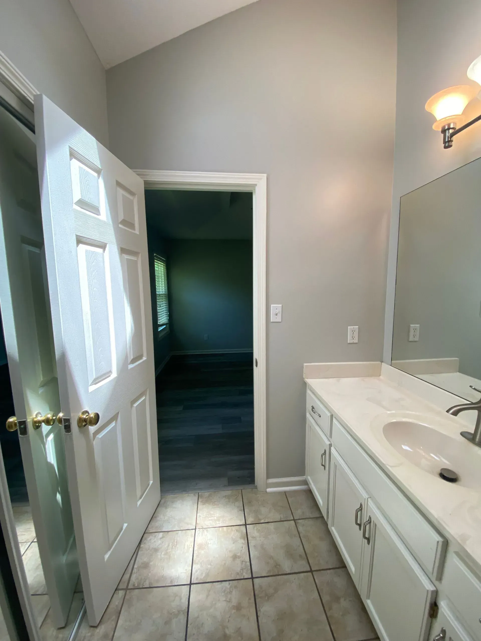 Bathroom with white vanity, open door to bedroom, tile floor, gray walls.