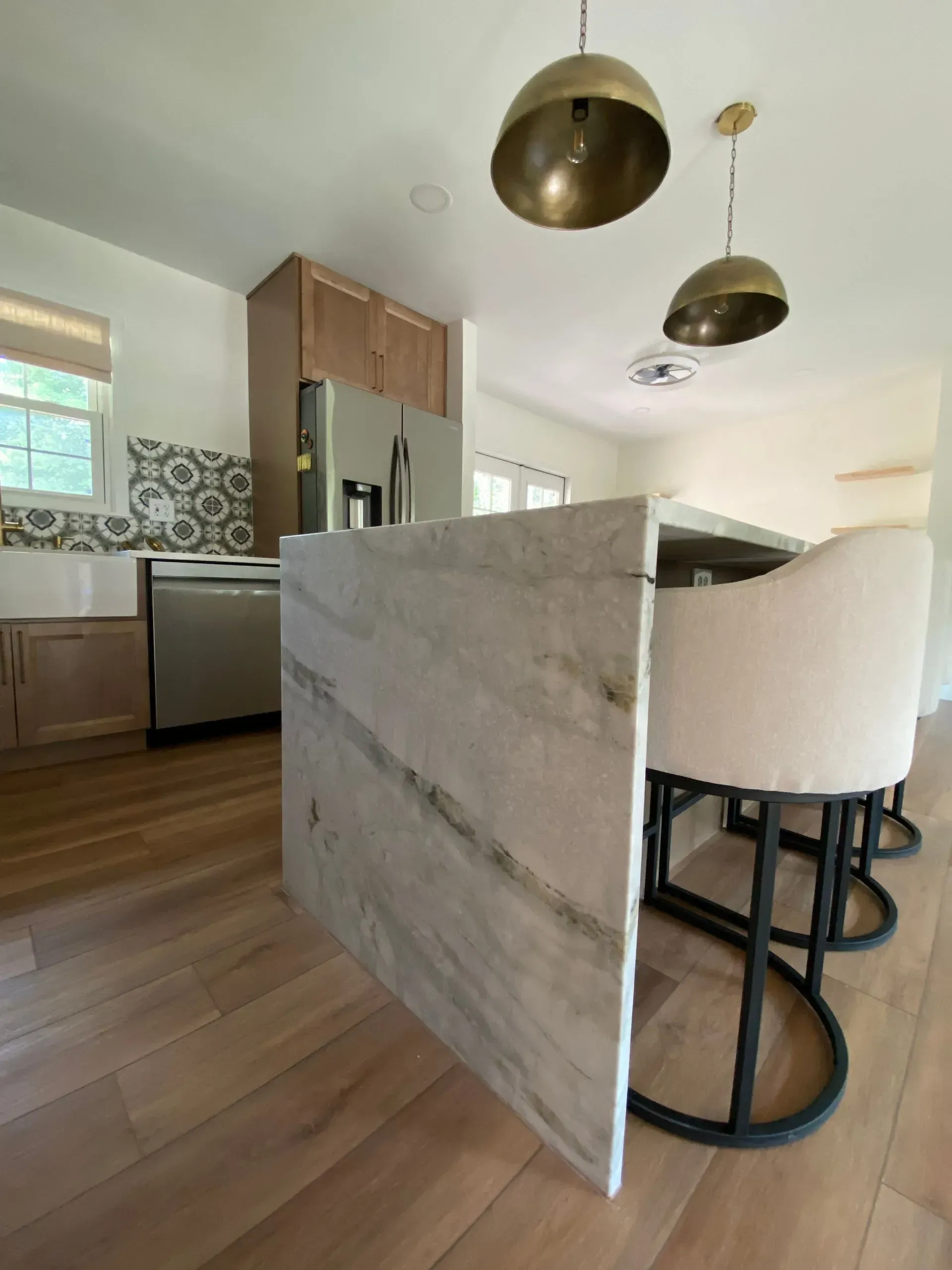 Kitchen with marble island, brass pendant lights, light wood cabinets, stainless steel appliances, and beige bar stools.
