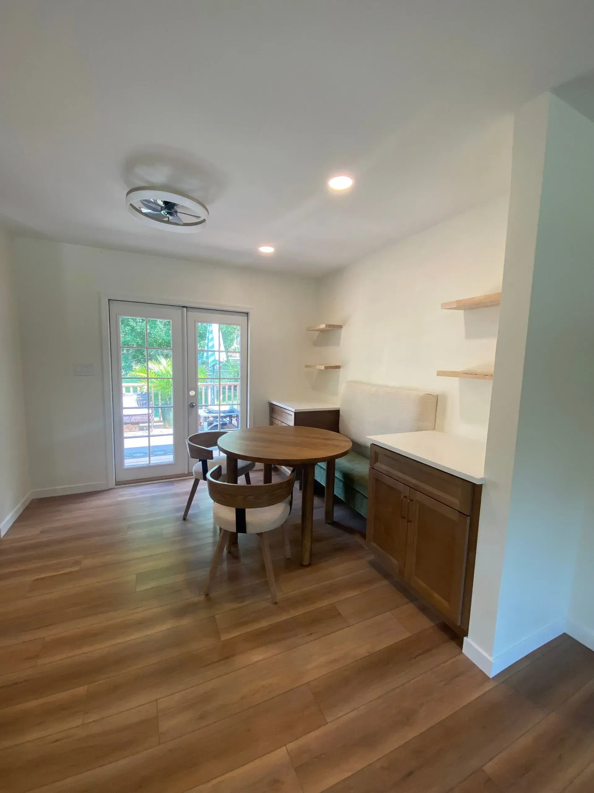 Dining area with wooden floor, table and chairs, built-in bench, and shelves. Doors open to outside.