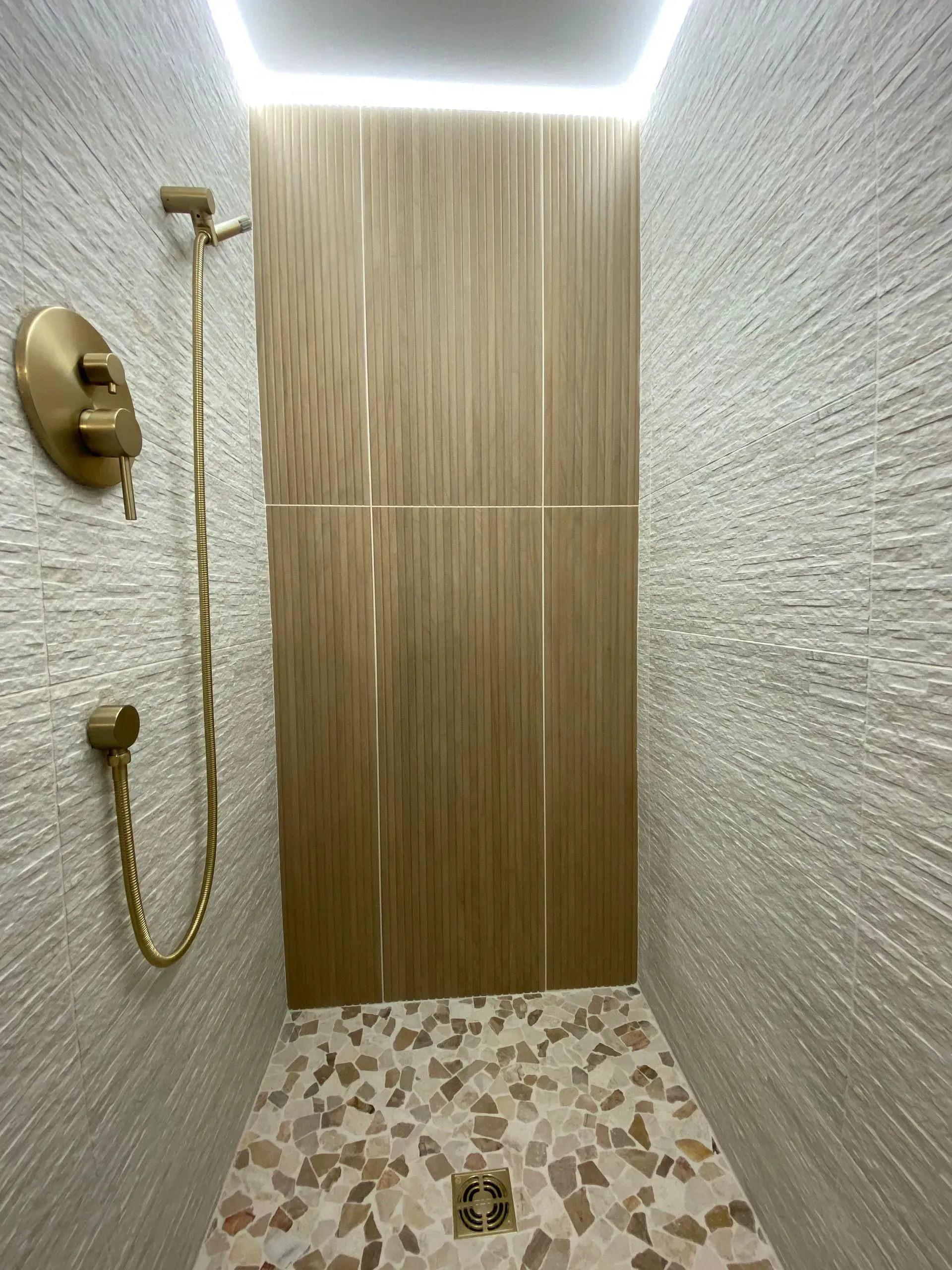 Shower with textured walls, wood-look center panel, and pebble floor. Gold shower fixtures.