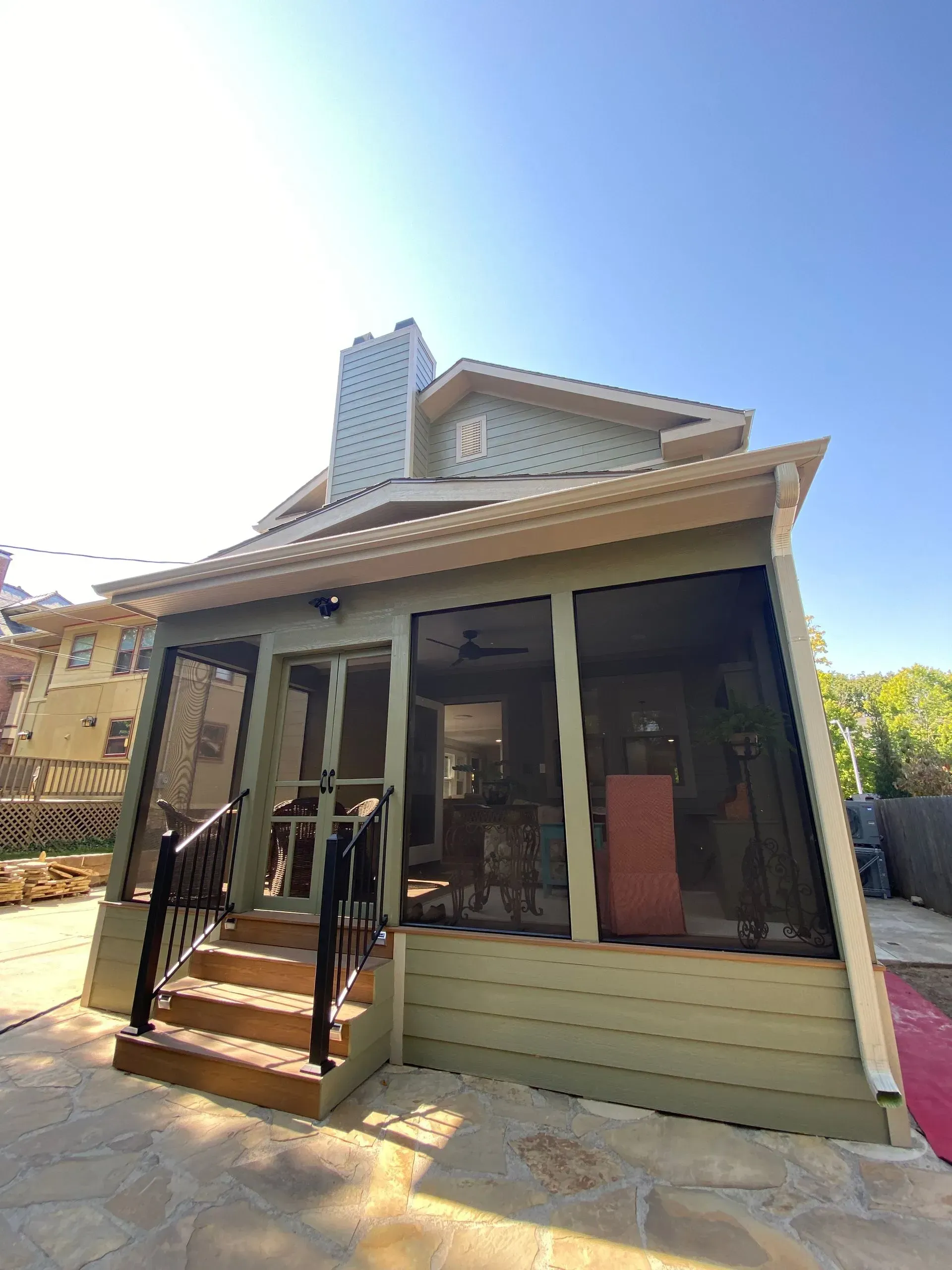 Screened-in porch on a two-story house with green siding, stairs, and black railing; chimney in the background.