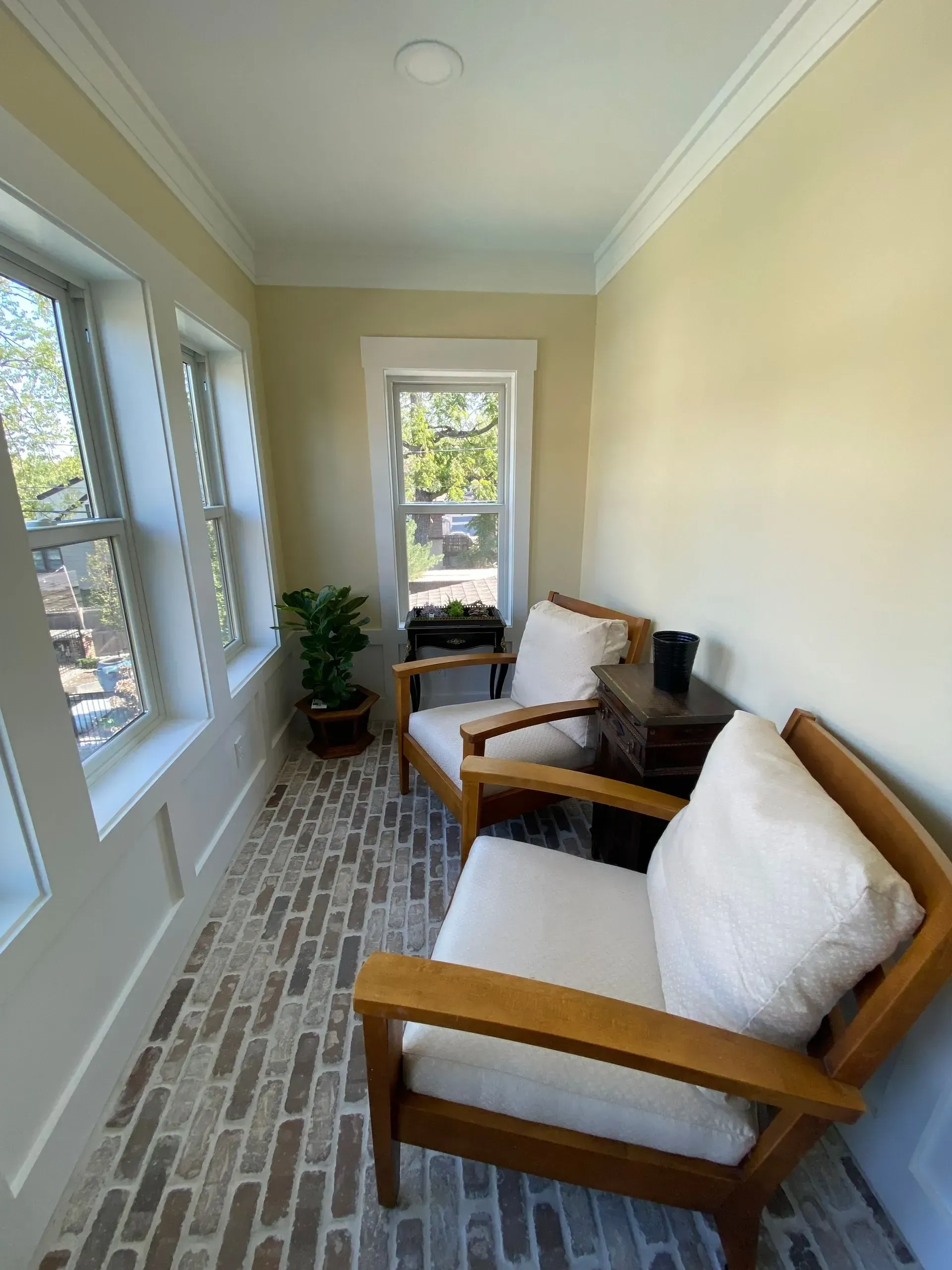 Sunroom with two wooden chairs, white cushions, small table, brick-like floor, windows.