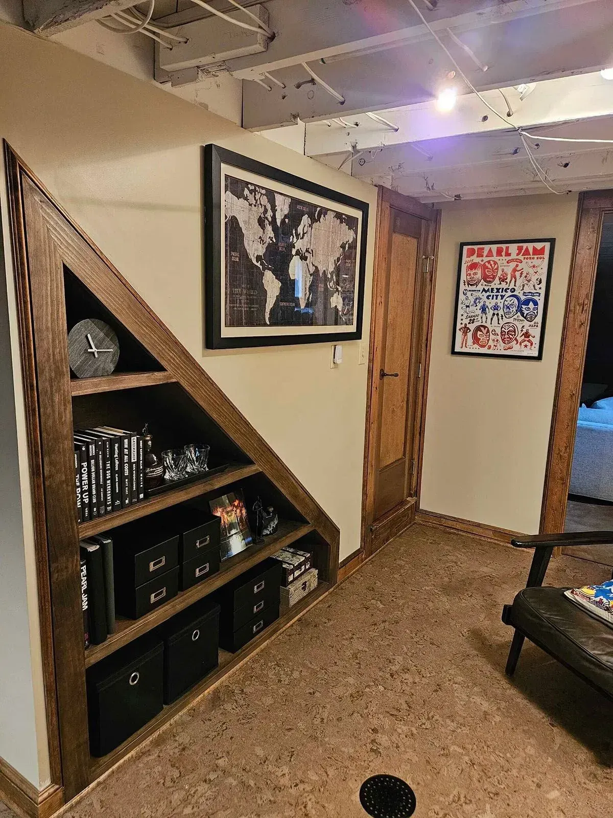 Built-in shelves under stairs, holding items, with a framed map and poster on the wall; cork flooring.