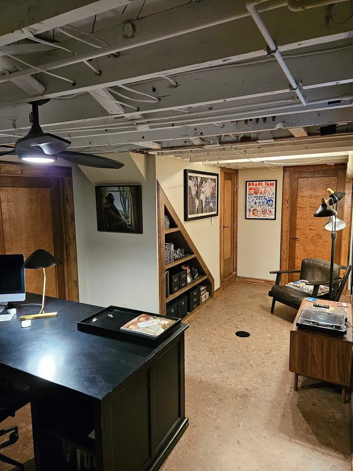Basement office with desk, bookshelves, framed art, ceiling fan, and arm chair.