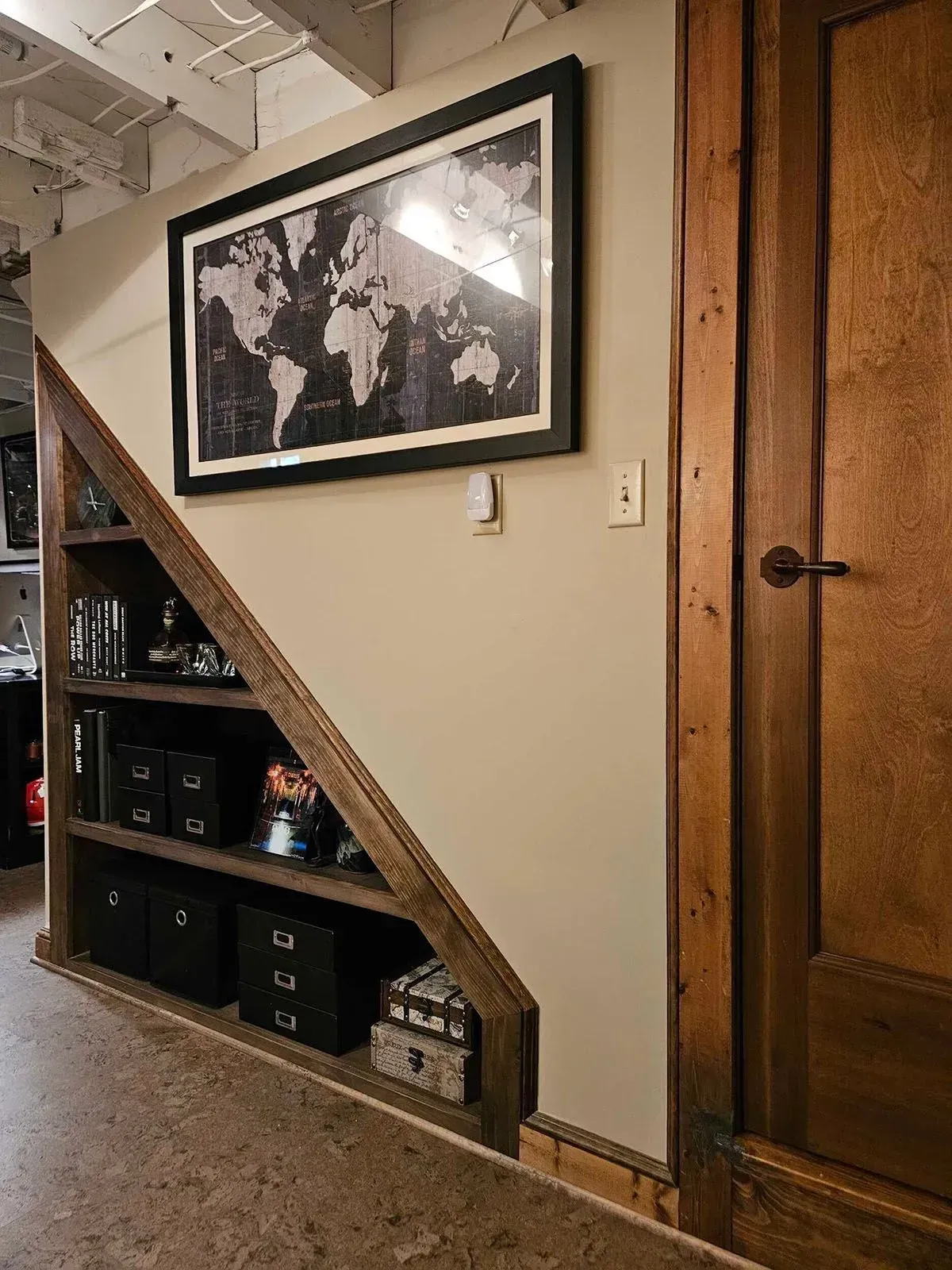 Built-in wooden bookshelf under stairs with framed world map and door on a neutral wall.