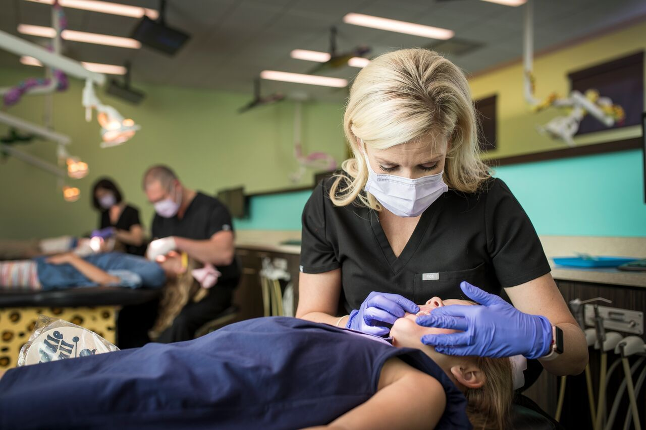 Dentist examining a young girl's teeth in a colorful dental office. Other dentists are treating patients in the background.