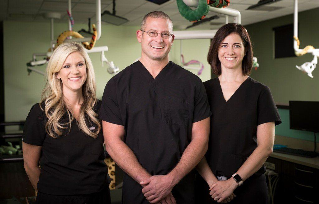 Three medical professionals in black scrubs smile in an operating room setting.