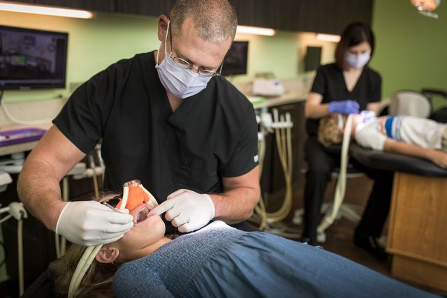 Dentist examining a child's teeth in an office, assistant in background.