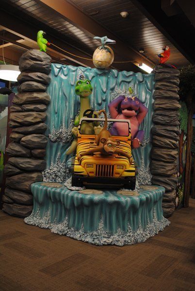 Dinosaur jeep in front of a waterfall display, with rock pillars, colorful cartoon dinosaurs and a tropical setting.