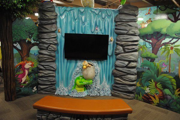 Jungle-themed play area: waterfall backdrop with TV, crocodile statue, orange bench, and jungle murals.