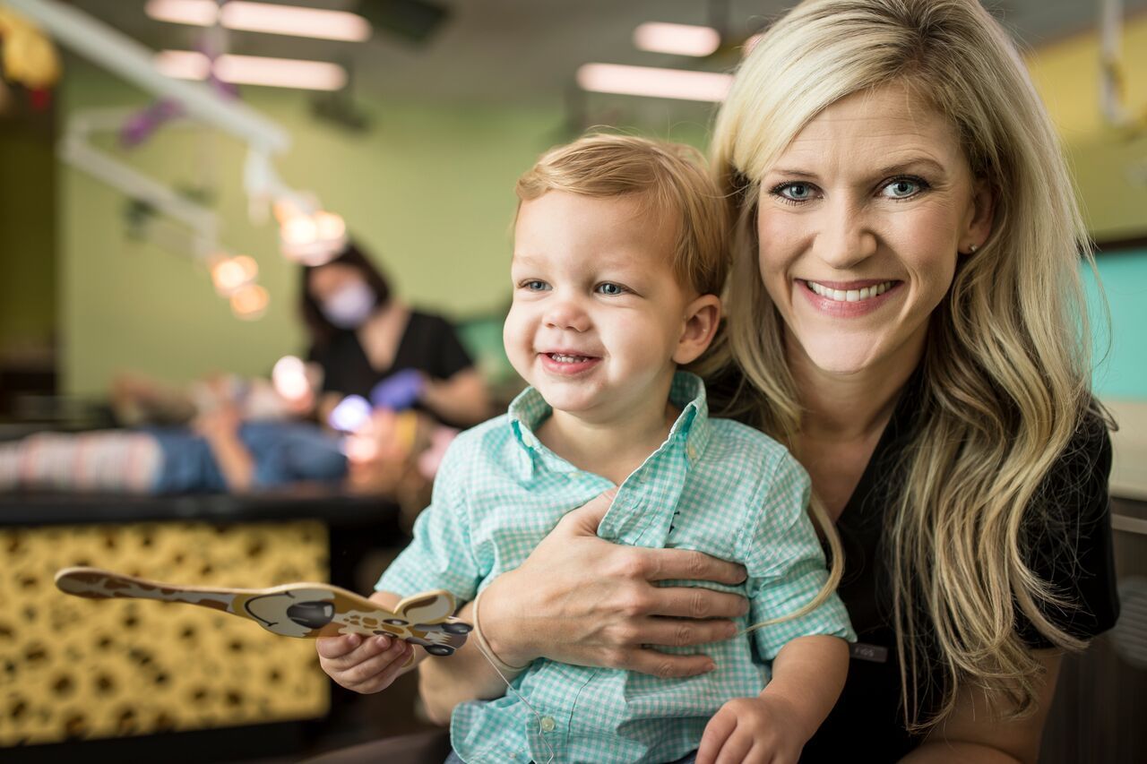 Smiling woman holding a toddler; they are at a dentist office; another child gets dental work.